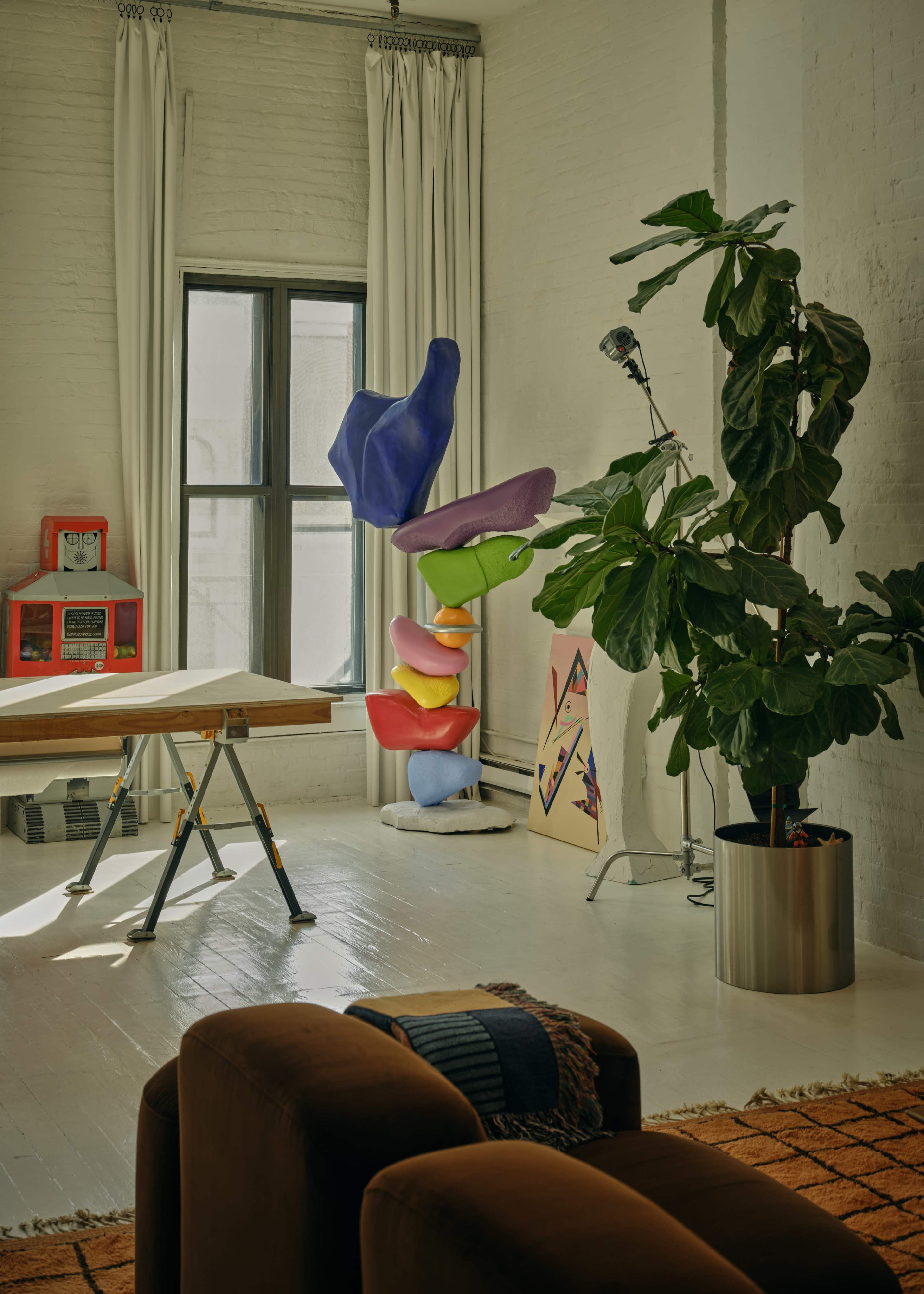 The image features a bright, modern interior with a colorful abstract sculpture, a potted plant, a drafting table, and an artwork leaning against the wall.