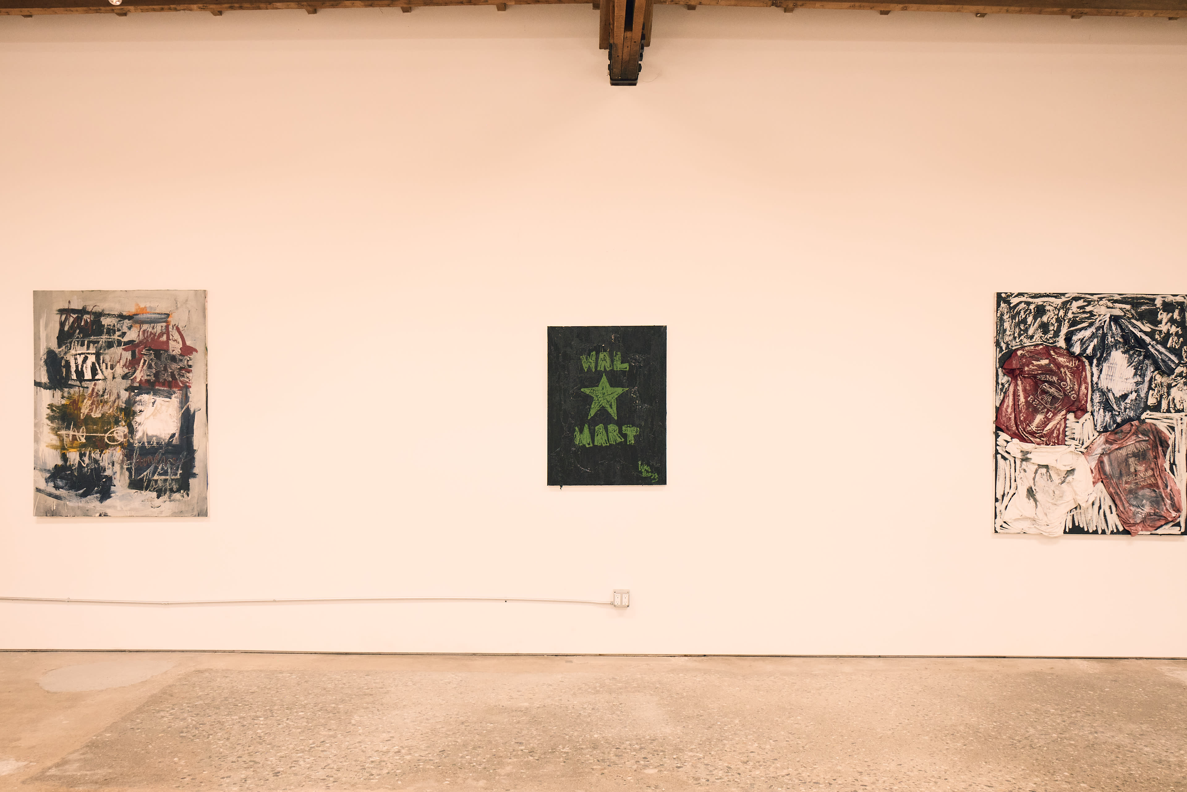 Three abstract paintings are displayed on a white wall in a gallery setting, with one centered and featuring green text.