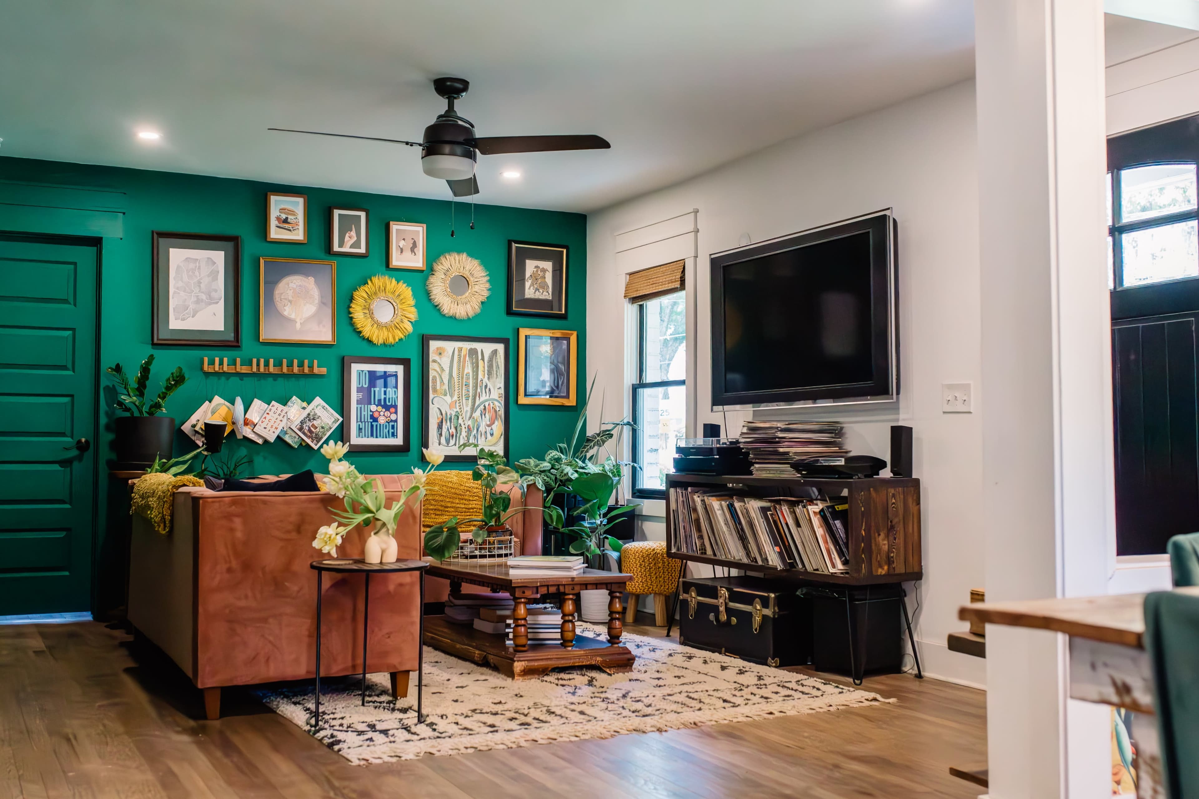 The living room features a green accent wall, a brown sofa, a collection of framed art, a television, and a wooden coffee table surrounded by plants.