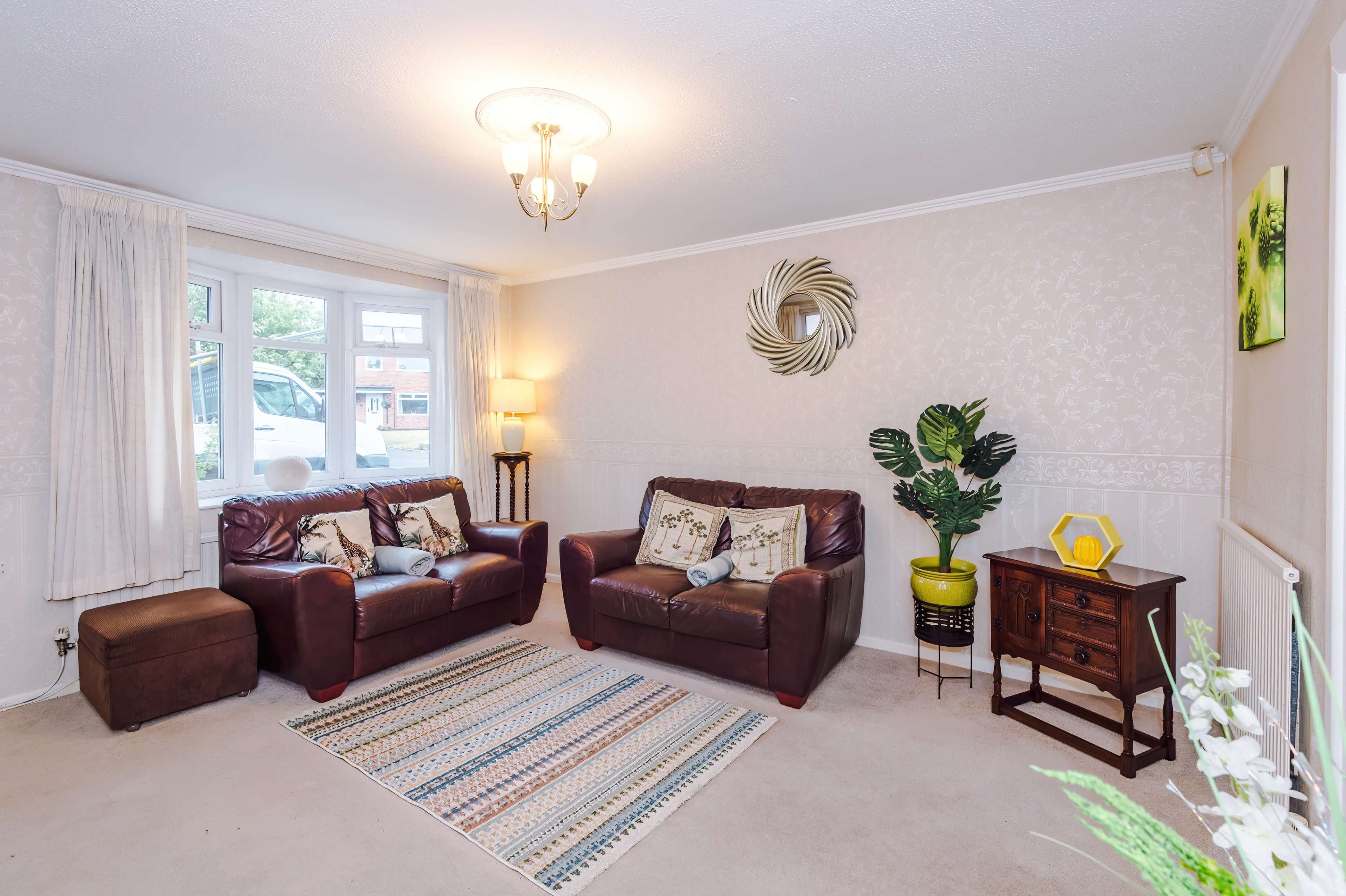 The image shows a living room with two brown leather sofas, a small wooden table, a potted plant, and a patterned rug, illuminated by a ceiling light.
