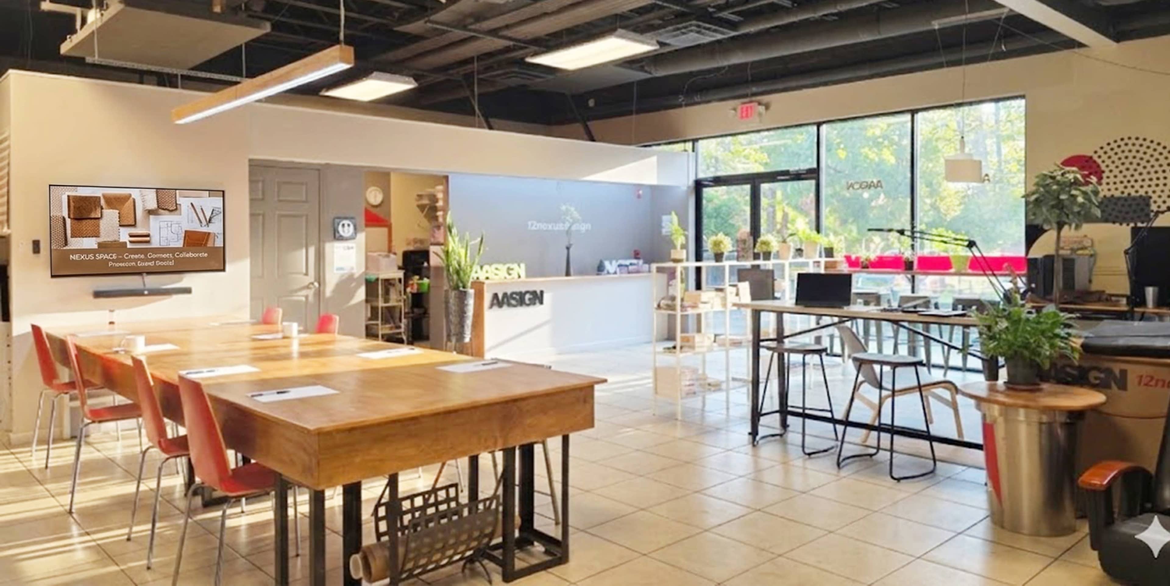 The image shows a modern, open interior workspace featuring a large wooden table surrounded by chairs, a reception area, and hanging lighting fixtures.