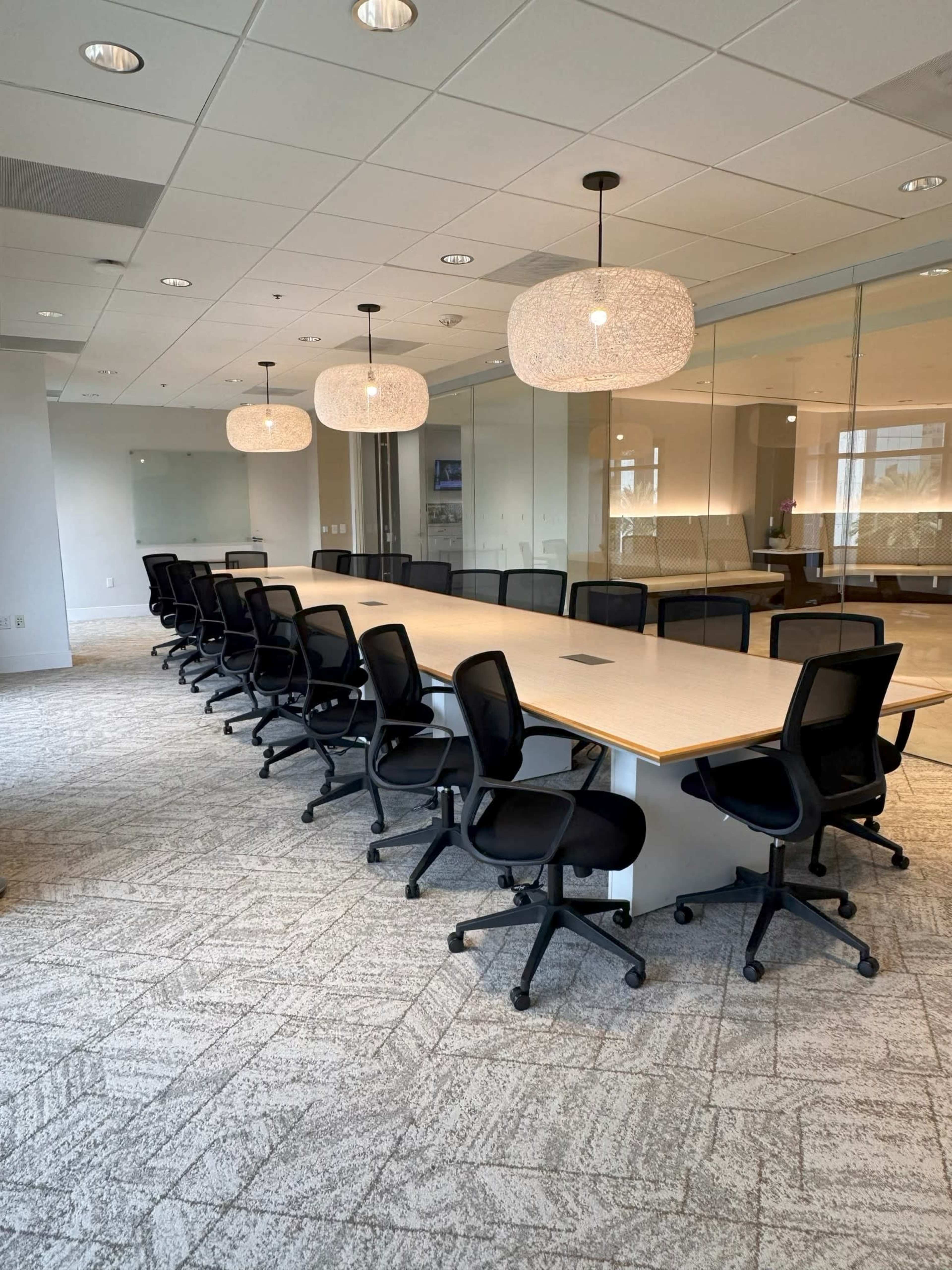 A long conference table with black chairs is set up in a well-lit meeting room featuring large windows and modern lighting fixtures.