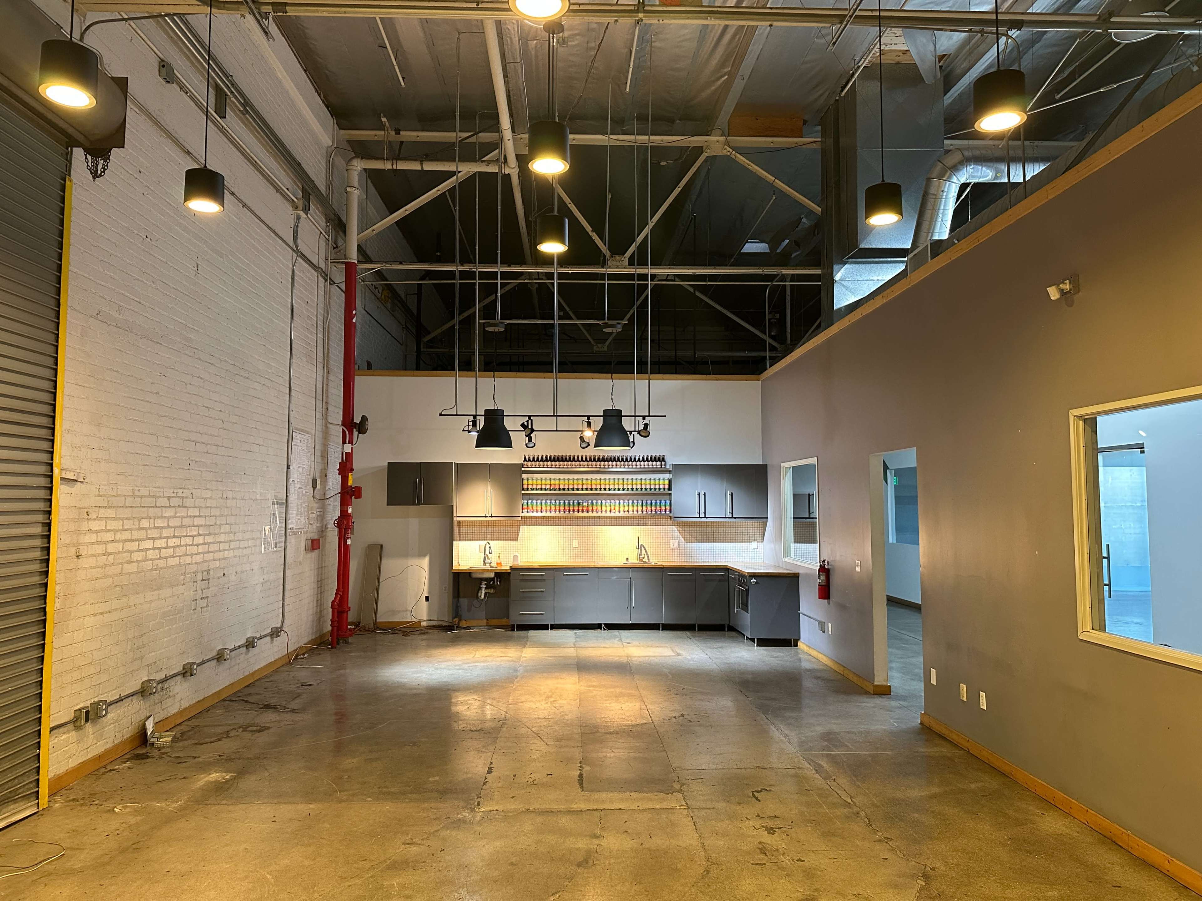 The image shows an industrial-style open space with high ceilings, concrete floors, and a kitchenette along one wall, illuminated by overhead lights.