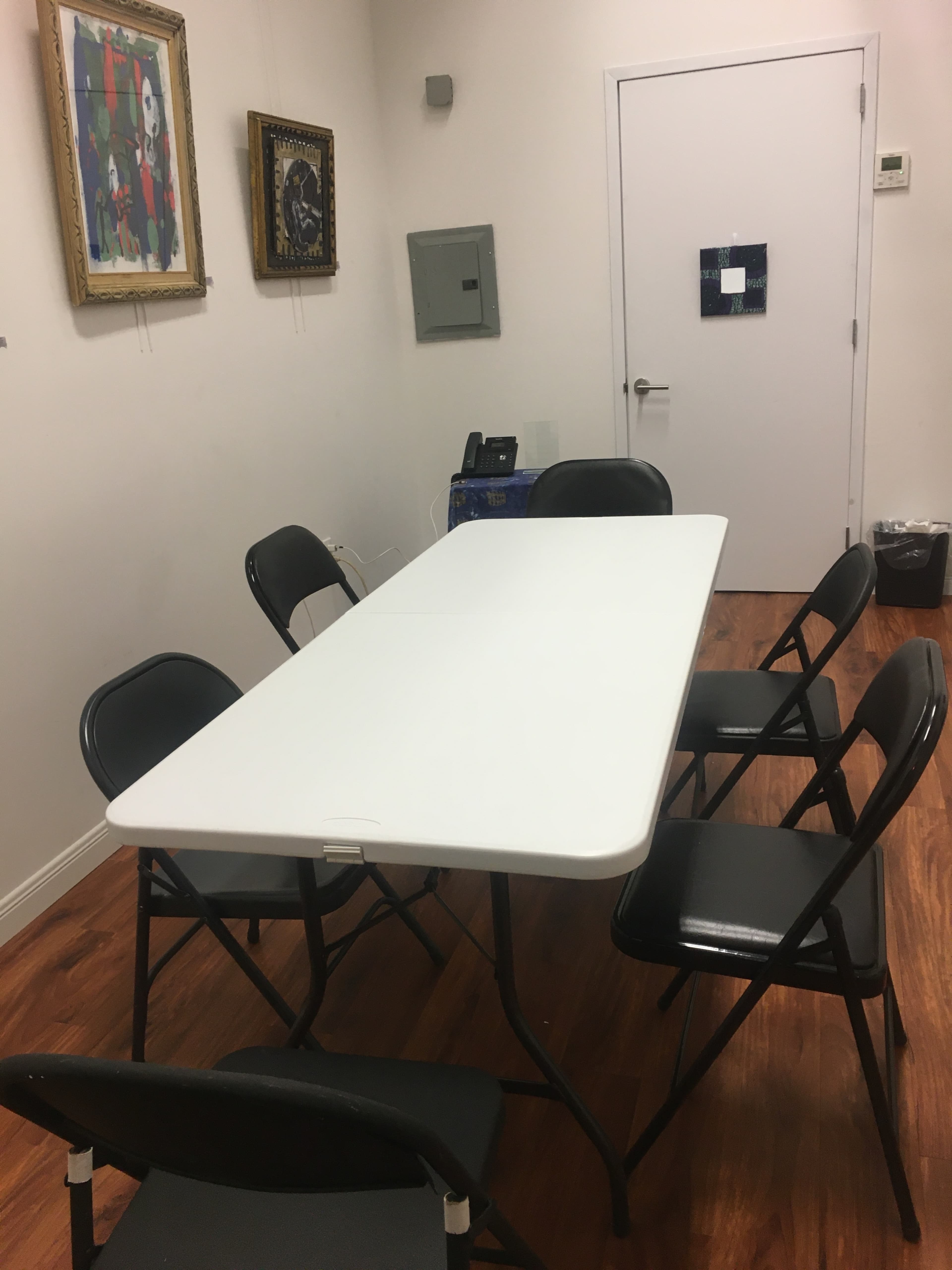 A white folding table is surrounded by six black folding chairs in a small, brightly lit room with framed artwork on the walls.