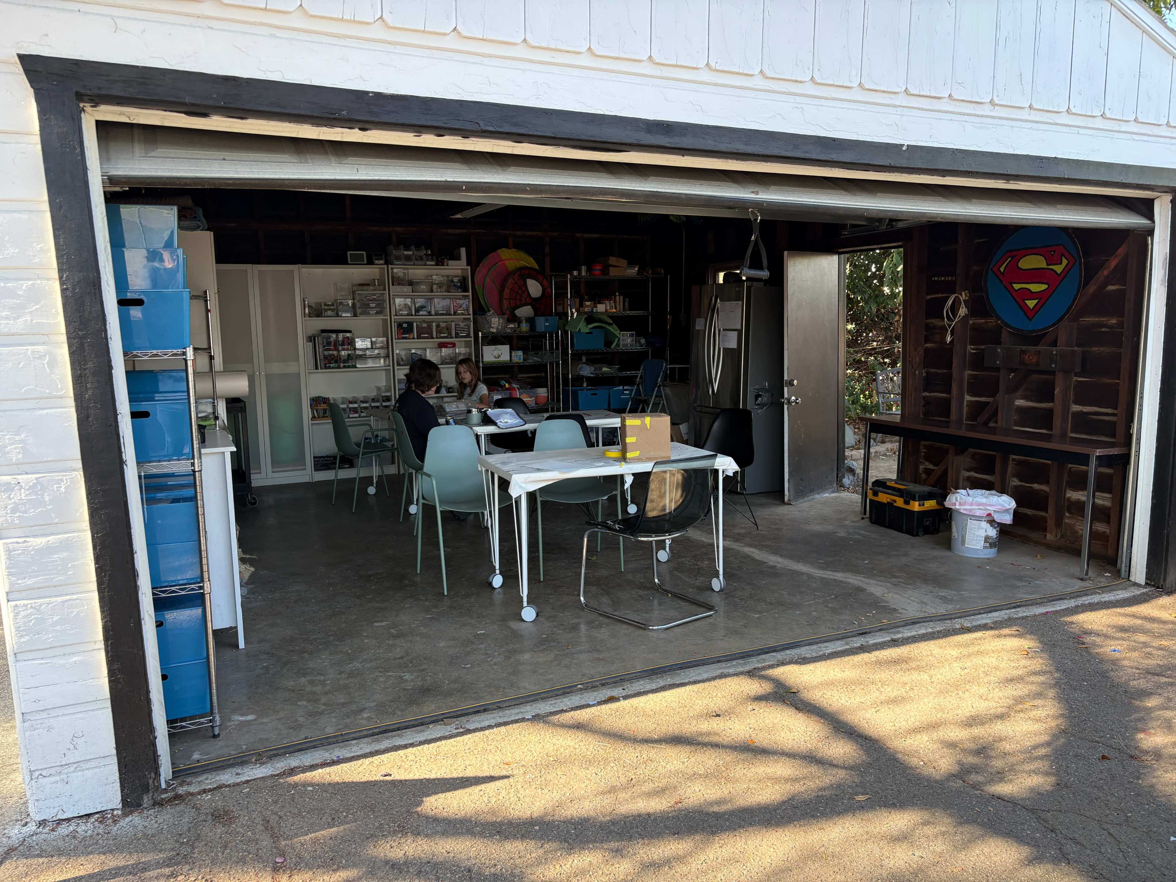 A garage with an open door features tables, chairs, and various storage items, while a large Superman emblem decorates the interior wall.