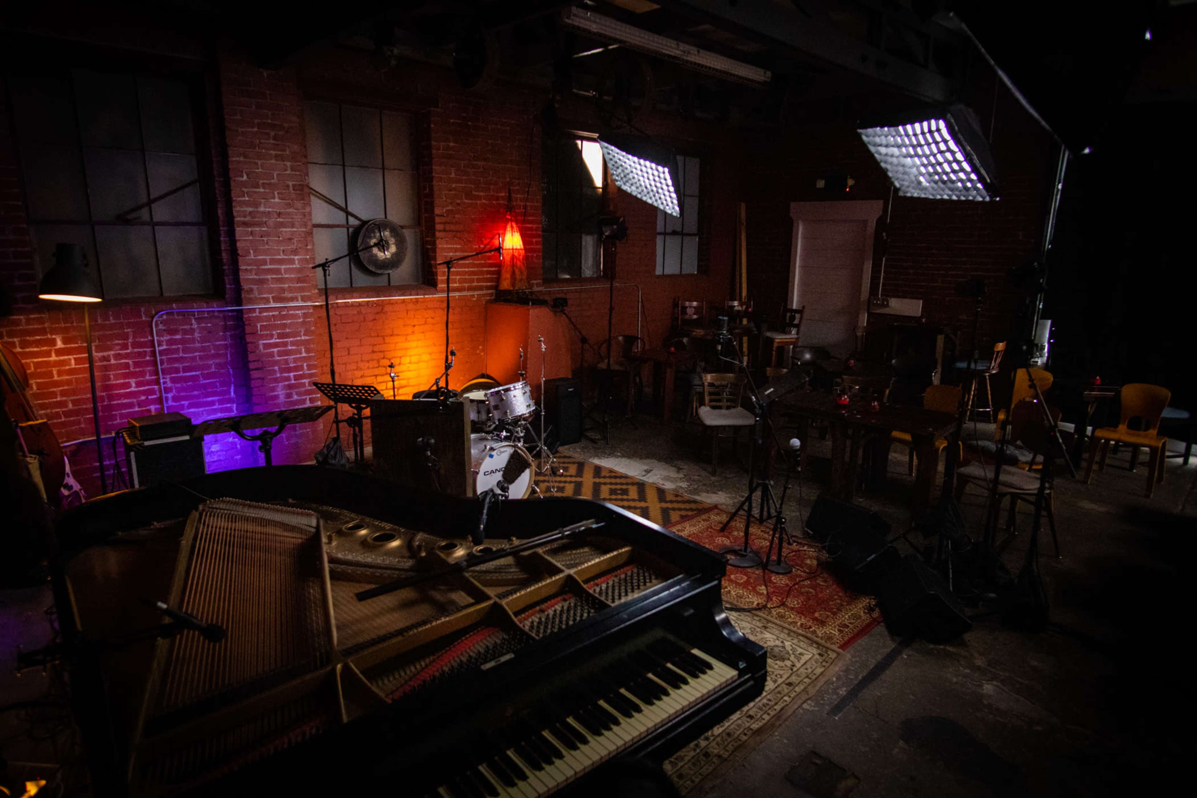 A dimly lit music venue with a grand piano in the foreground and various instruments set up, including a drum set and amplifiers against exposed brick walls.