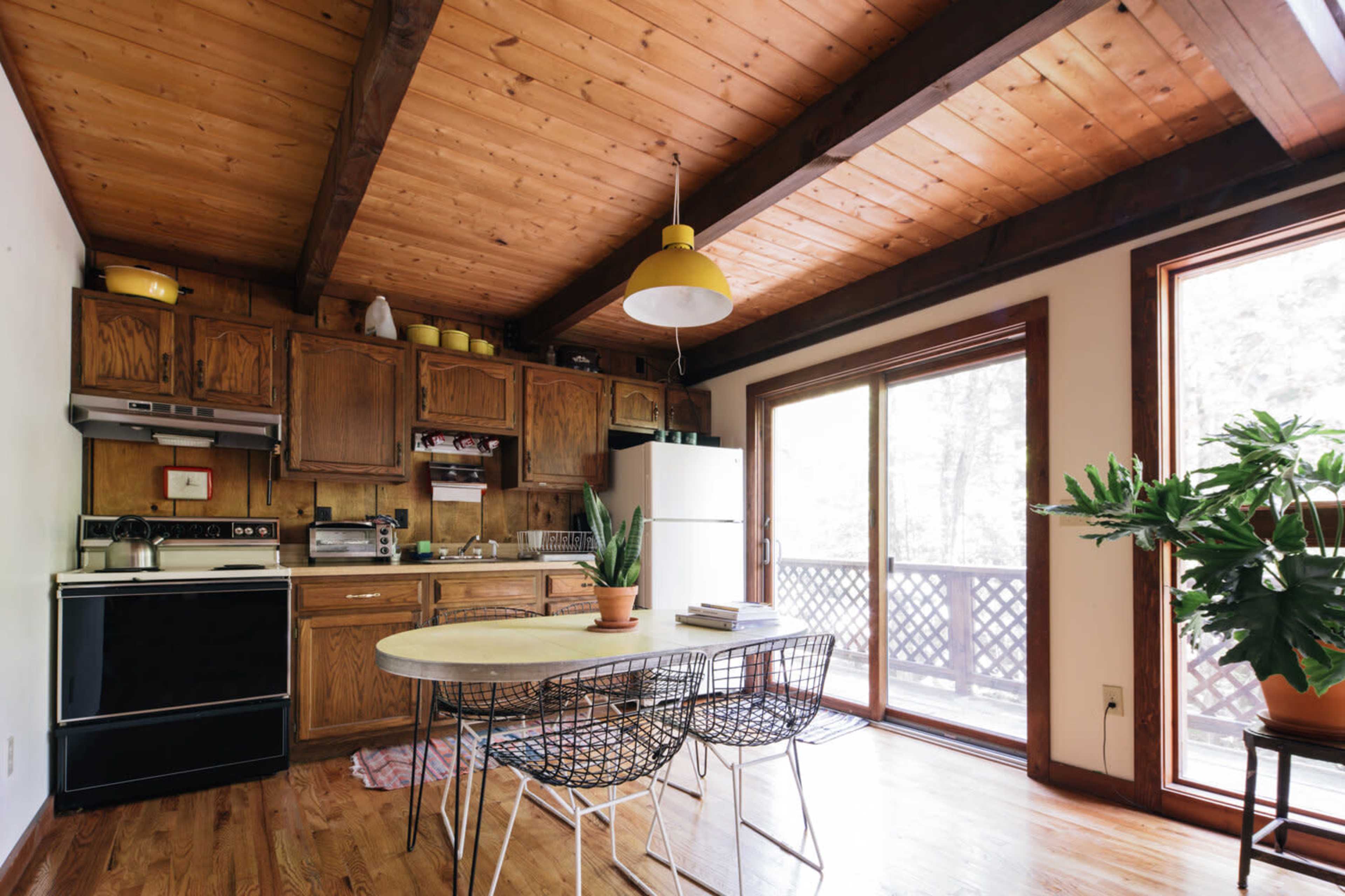 A wooden kitchen features brown cabinetry, a round dining table with wire chairs, and large windows overlooking a deck.