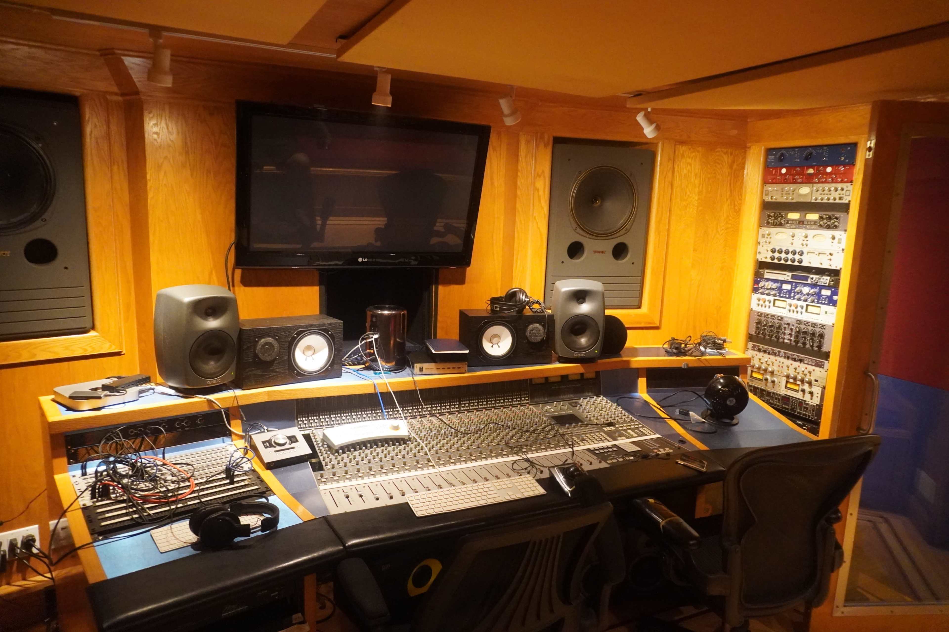 A recording studio with a large mixing console, multiple speakers, a television monitor, and various audio equipment mounted on the wall.