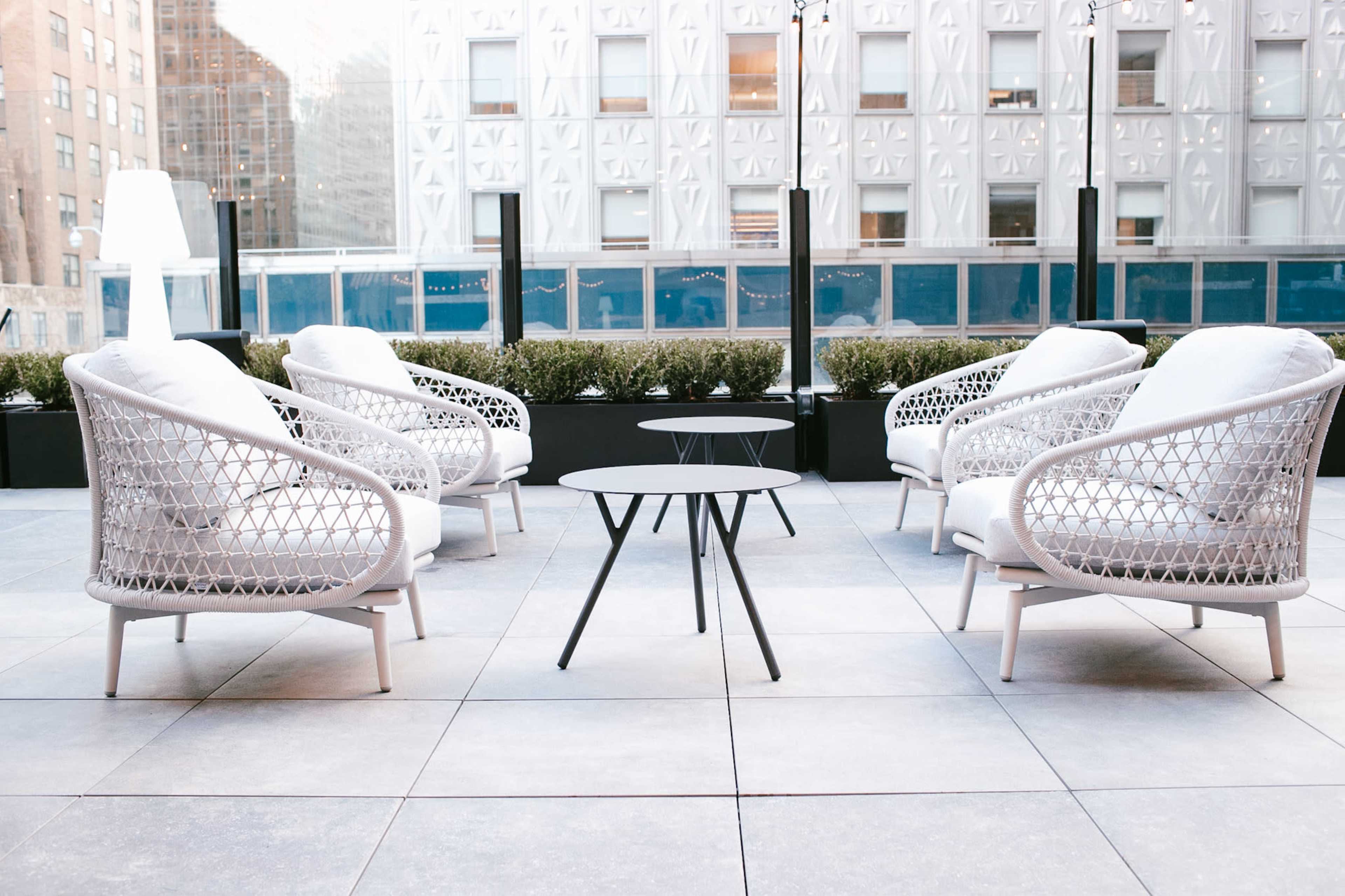 A seating area features four white, woven chairs arranged around a small black table on a patio with modern architecture in the background.