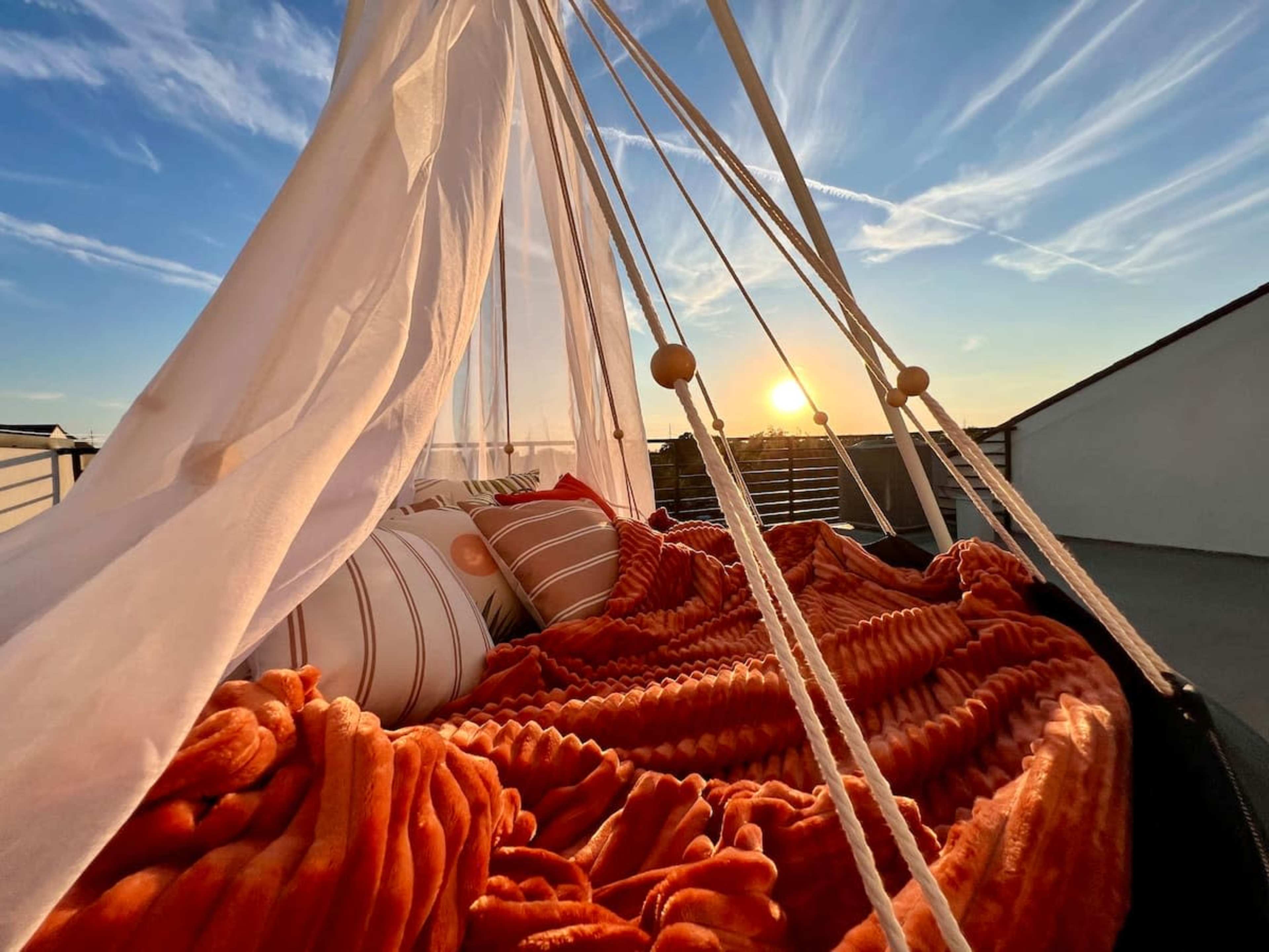 A cozy outdoor bed is draped with orange blankets and pillows, framed by a white canopy, with a sunset visible in the background.