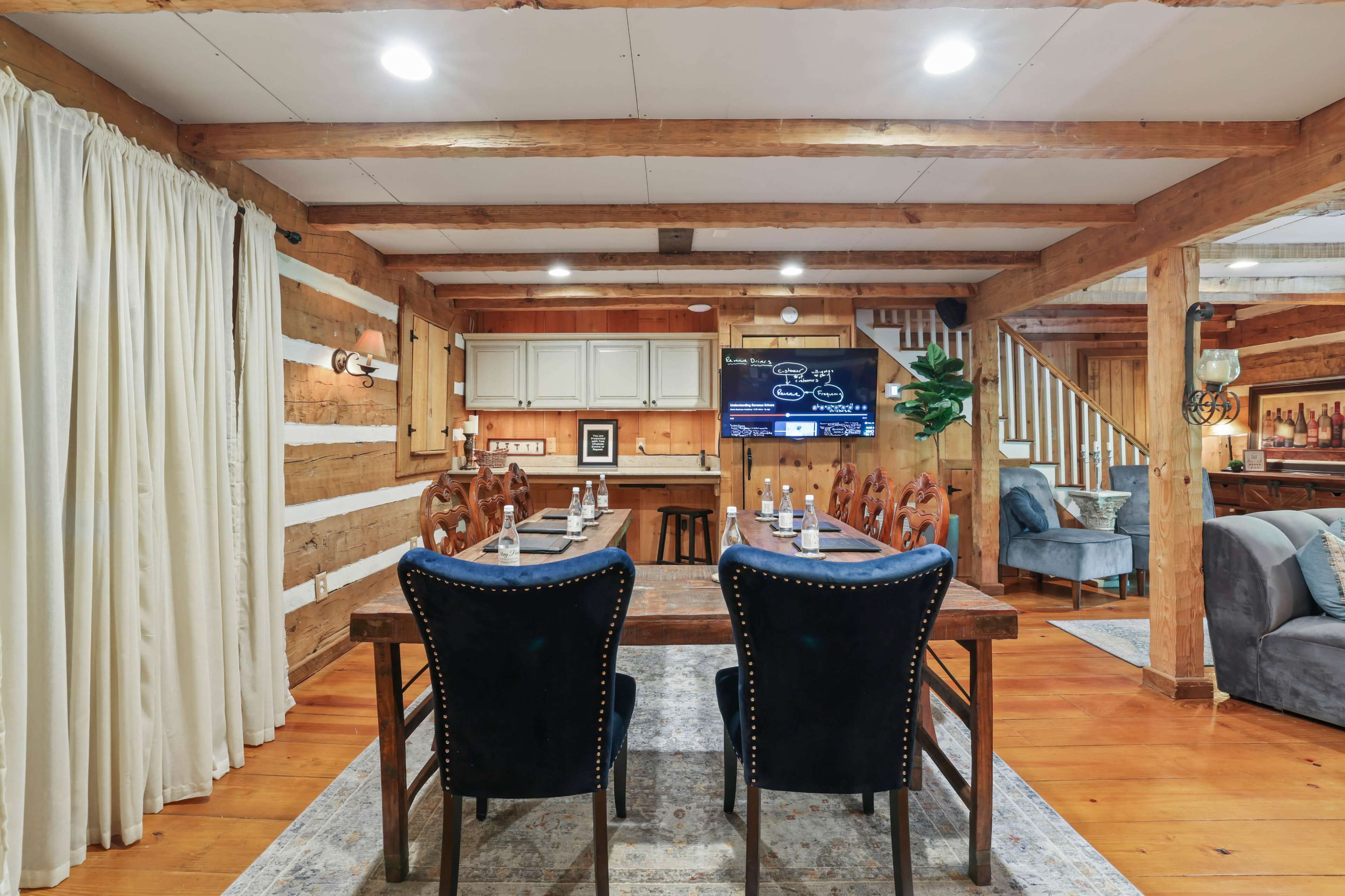 A wooden dining table with chairs is set in a rustic-themed room featuring exposed beams, a television, and a couch.