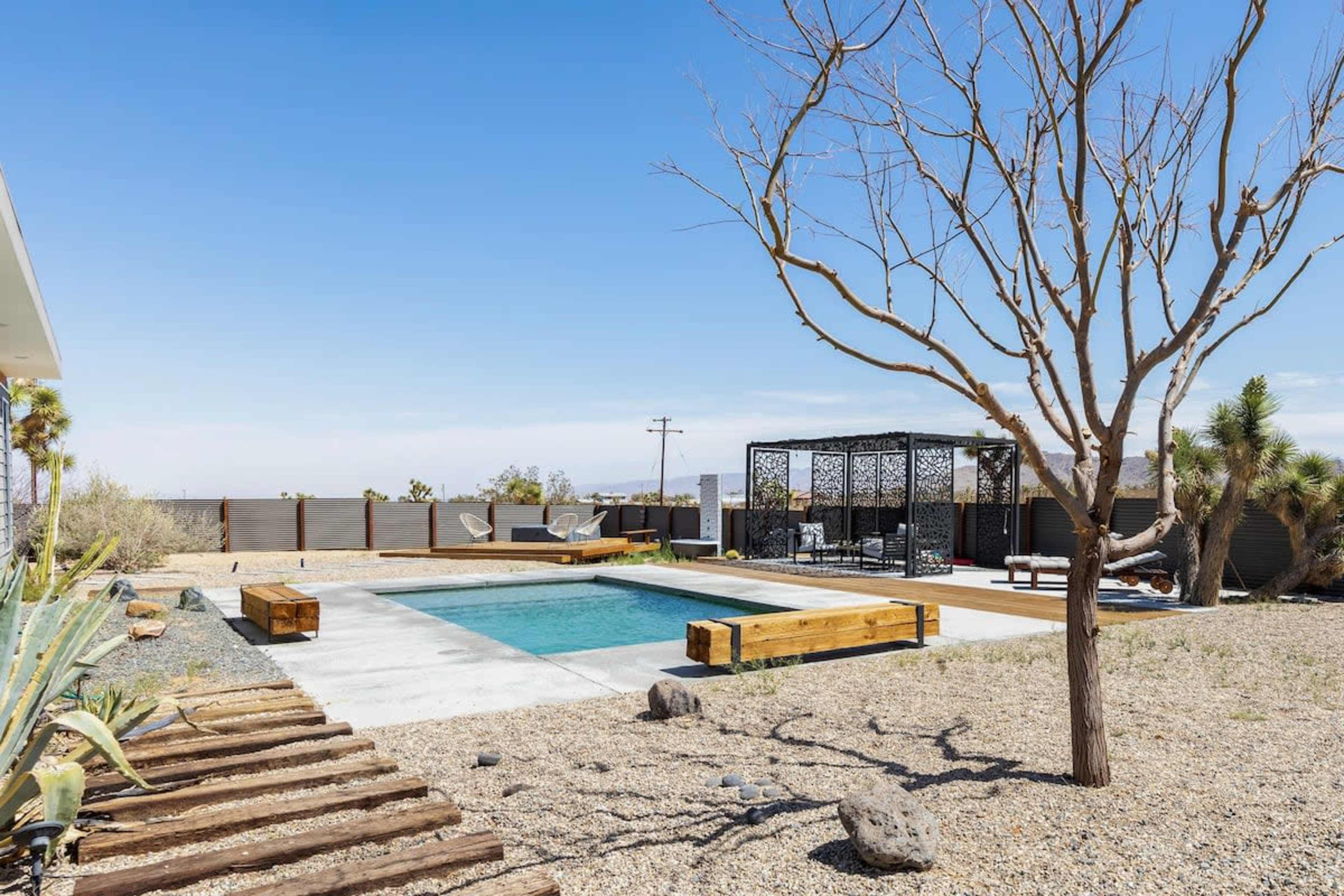 A spacious outdoor area features a rectangular pool surrounded by gravel, wooden seating, and a shaded gazebo, with a barren tree and desert vegetation nearby.