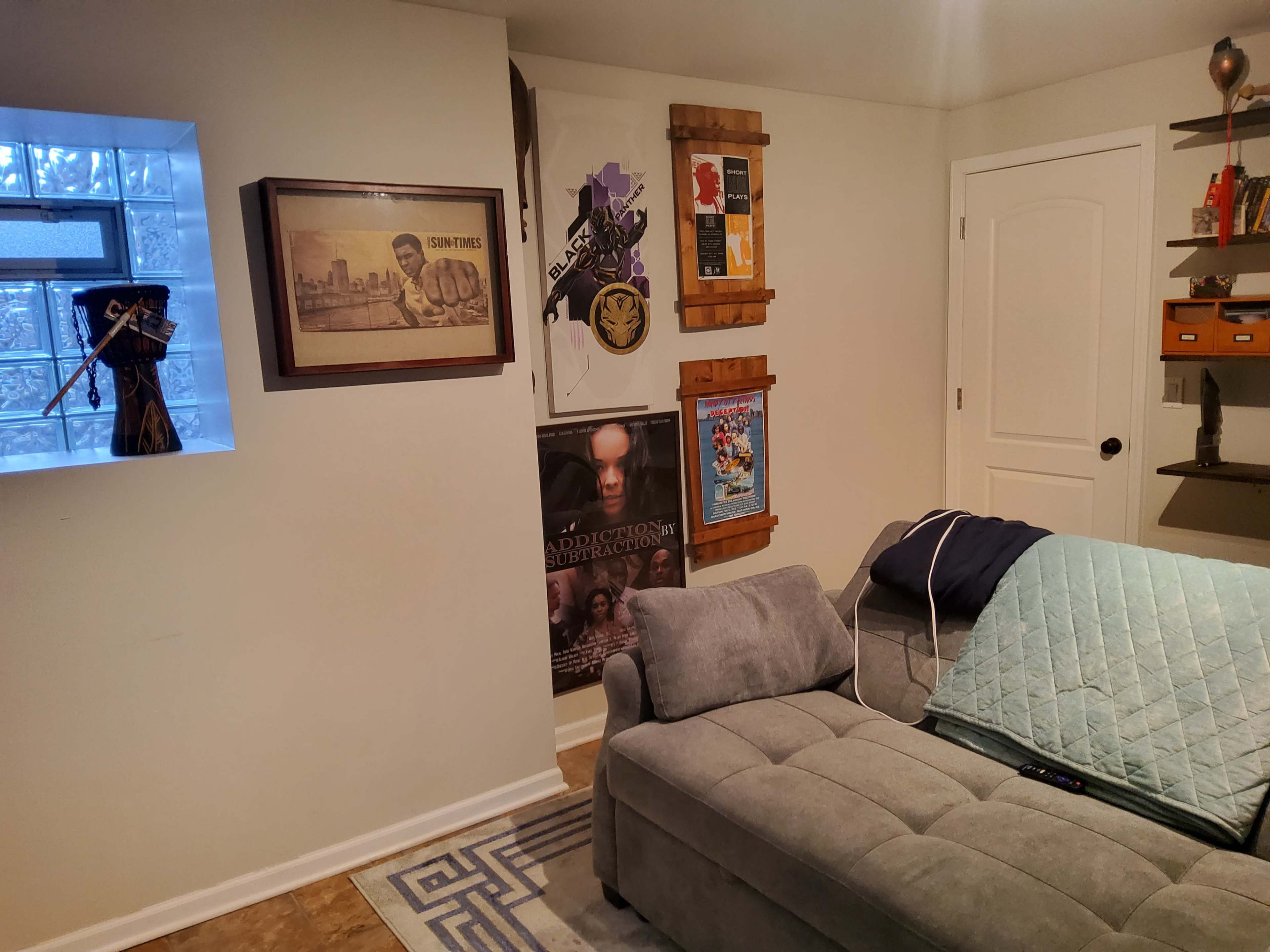 The image shows a cozy basement rec room with a gray sectional sofa, framed artwork on the wall, and an illuminated panel window.