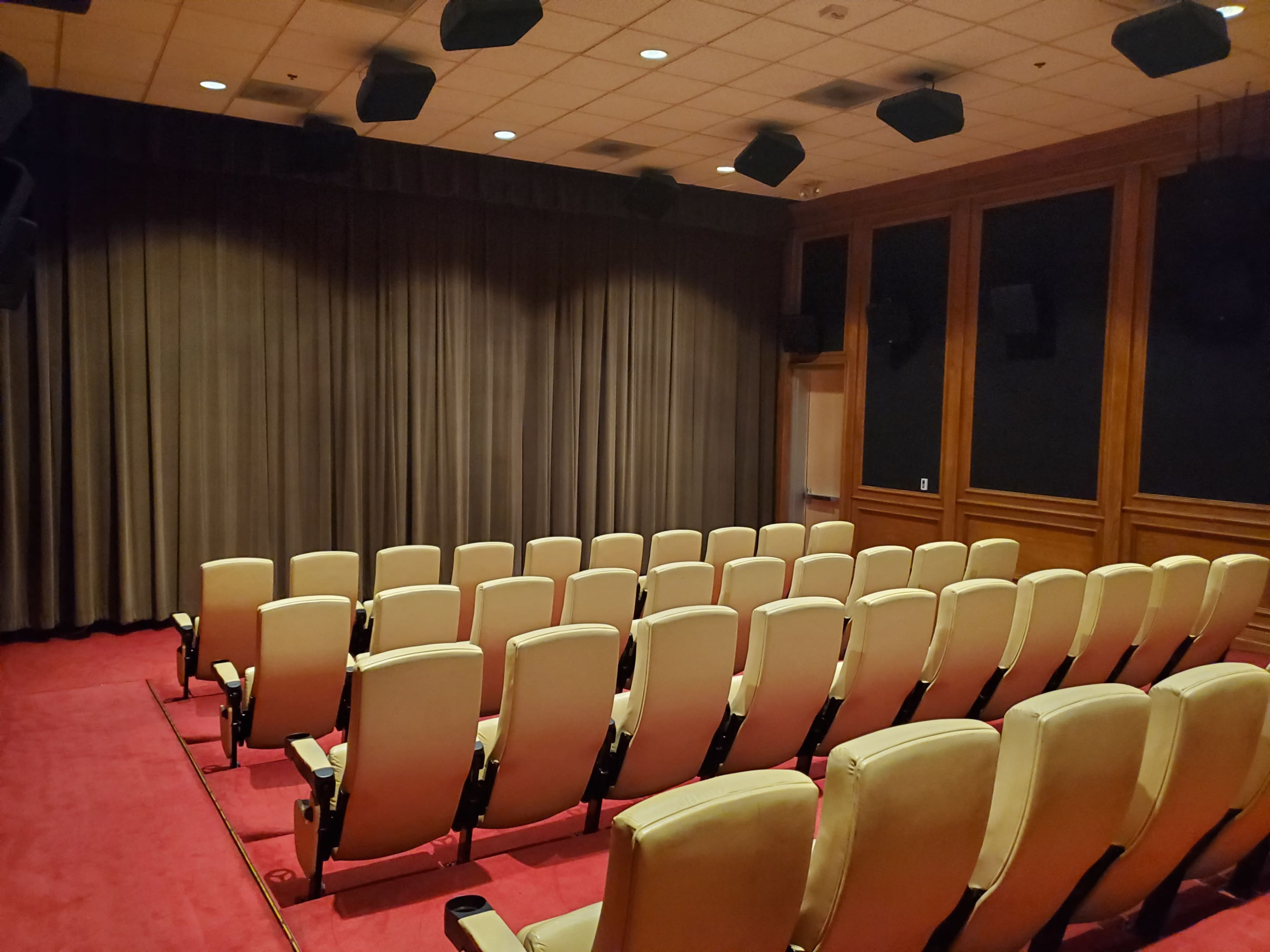 The image shows a small, empty theater with rows of cream-colored seats facing a closed curtain.