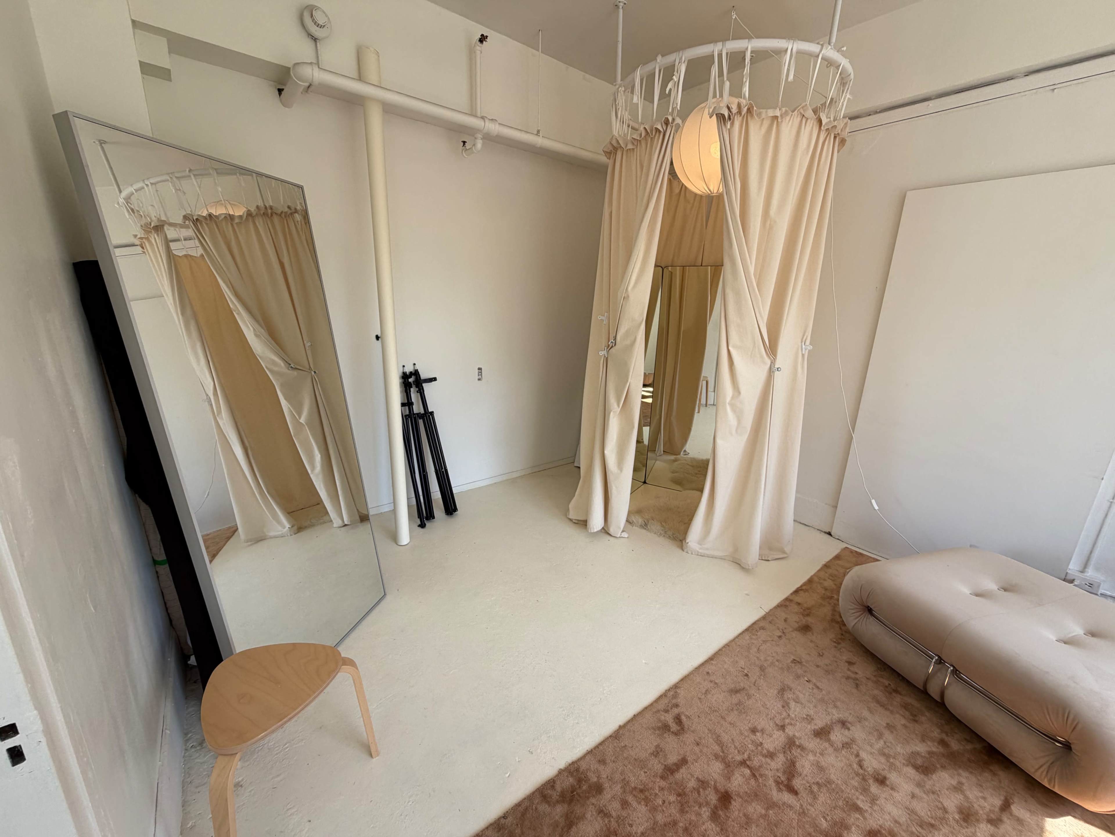 The image shows a minimalist room with a large mirror, a round curtain structure, a padded seat, and an area rug on a light-colored floor.