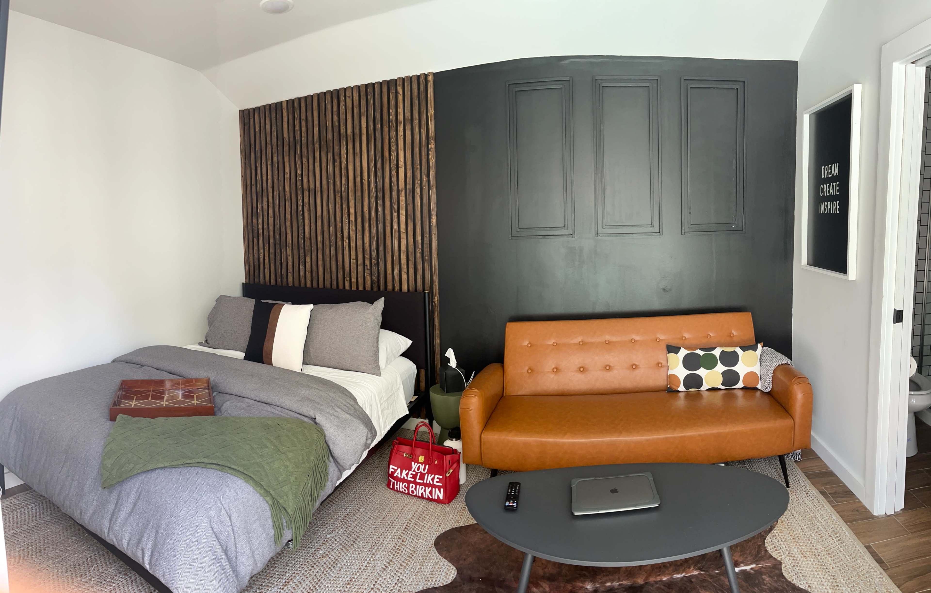 A small bedroom with a bed on one side and a brown couch with patterned cushions on the other, featuring a wall with wooden paneling and a dark painted section.