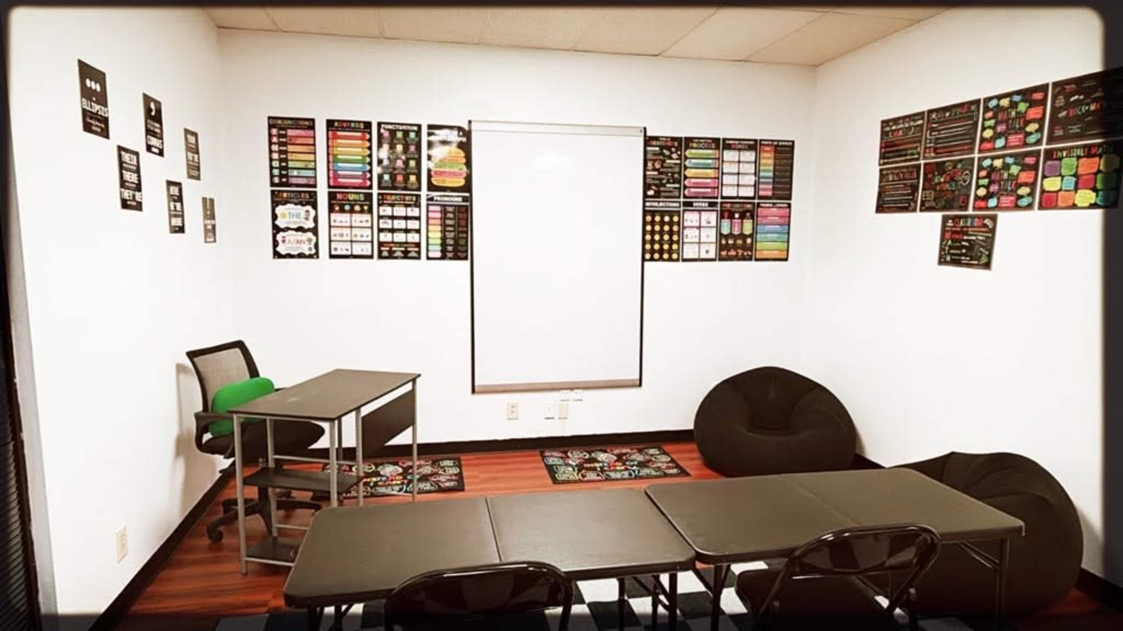 The image shows a classroom with two tables and chairs, bean bags in the corner, a whiteboard in the center, and various educational posters on the walls.