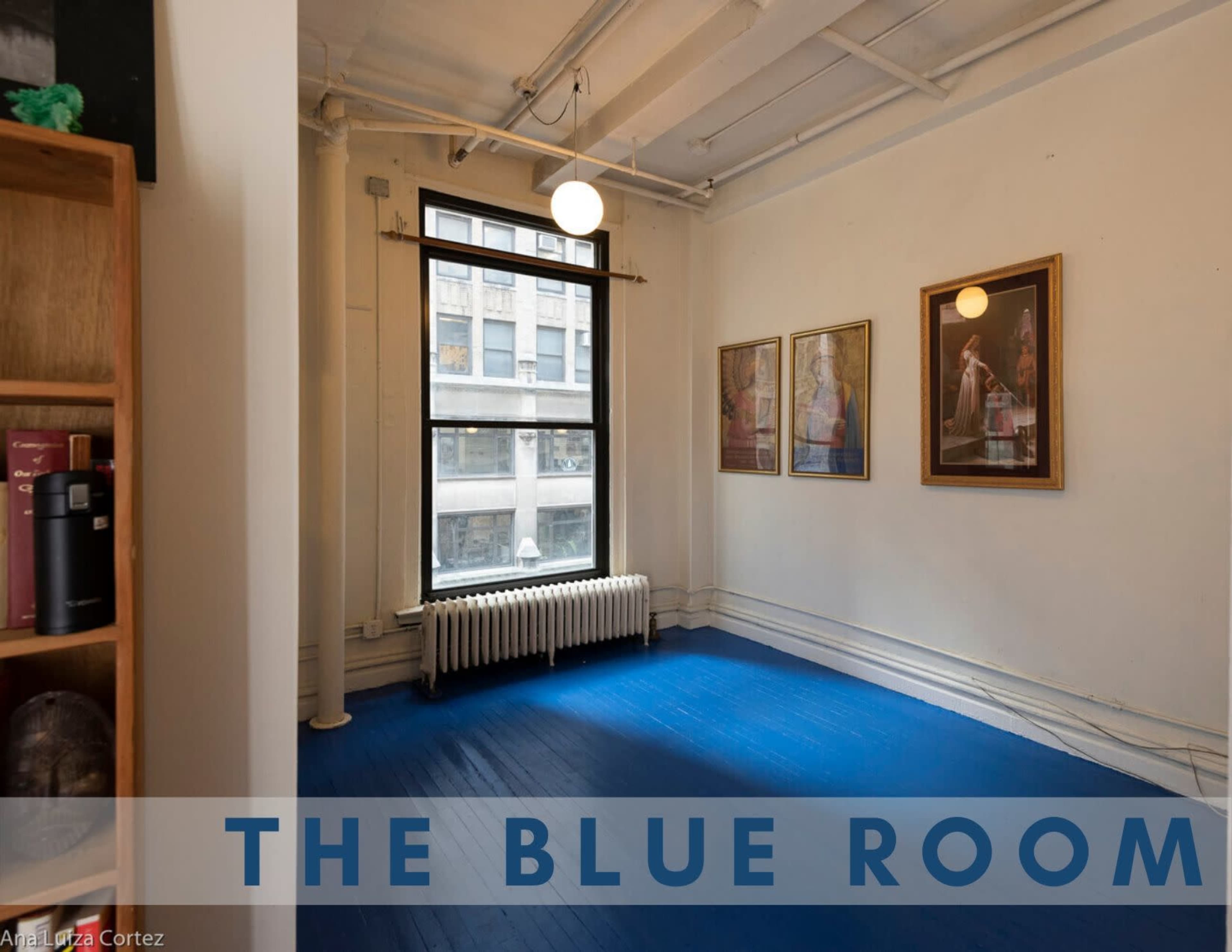 The image shows a room with blue flooring, large windows, and two framed artworks on the white walls.