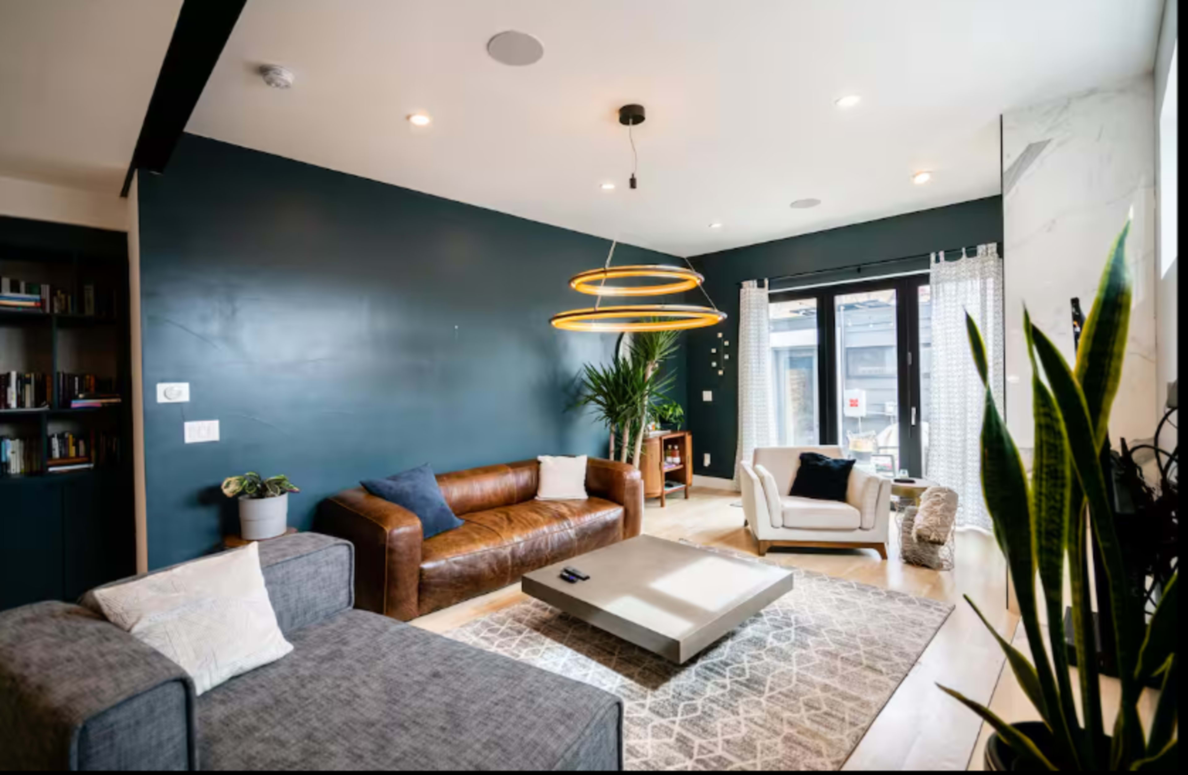 A modern living room features a brown leather sofa, a gray sectional, a round coffee table, and large windows with drapes, complemented by greenery and contemporary lighting fixtures.