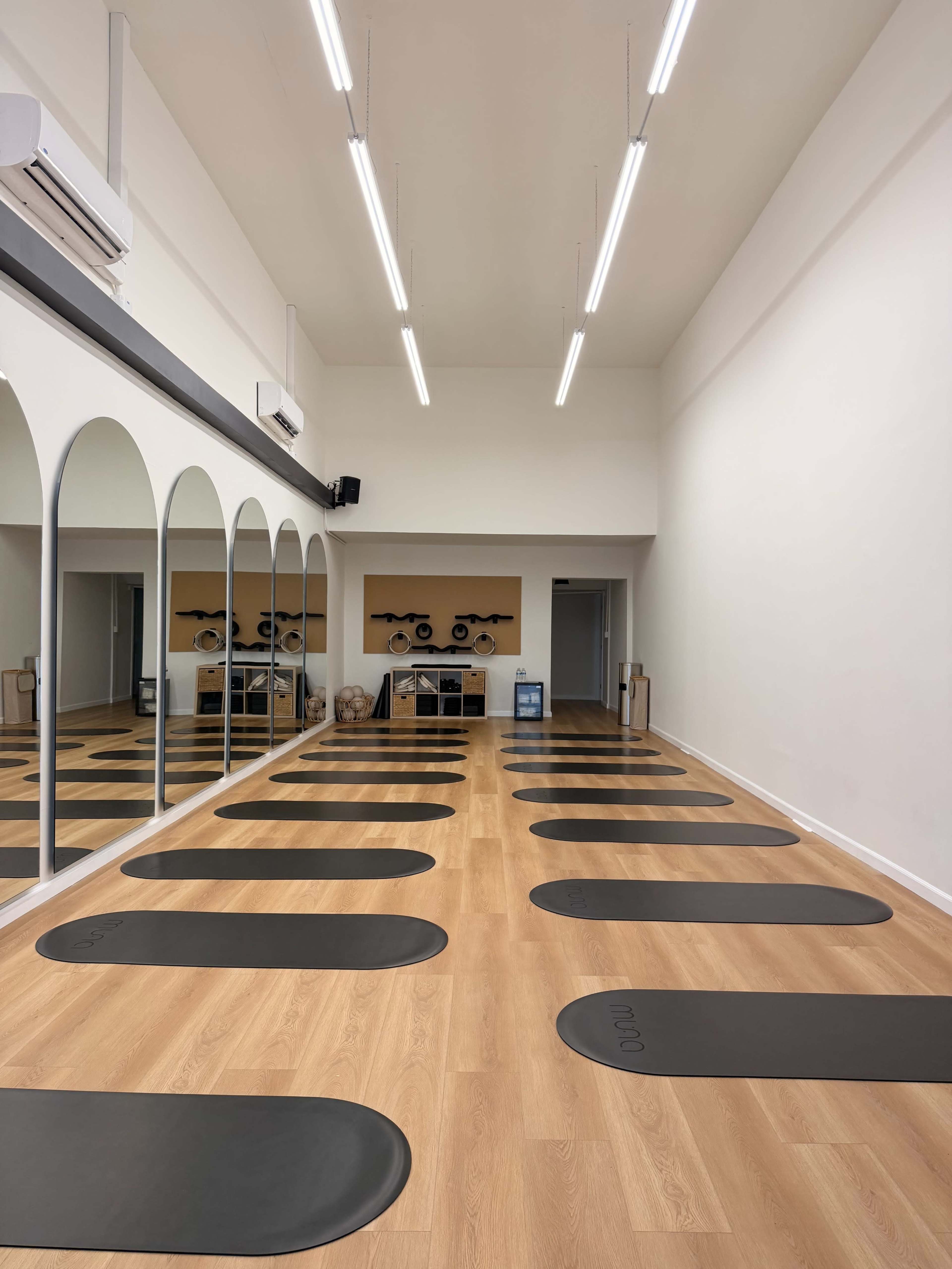 The image shows a spacious yoga studio with wooden floors, several yoga mats arranged in parallel, and large mirrors reflecting the room.
