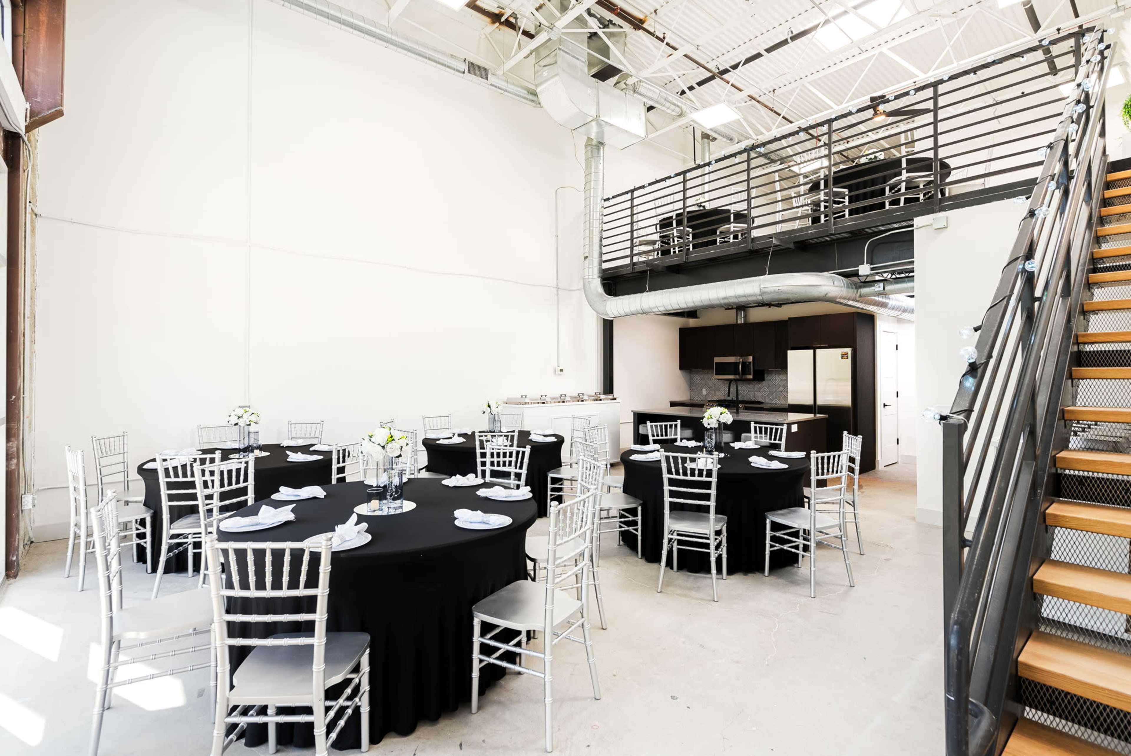 A spacious event venue with black round tables set for dining and a loft area overlooking the main floor.
