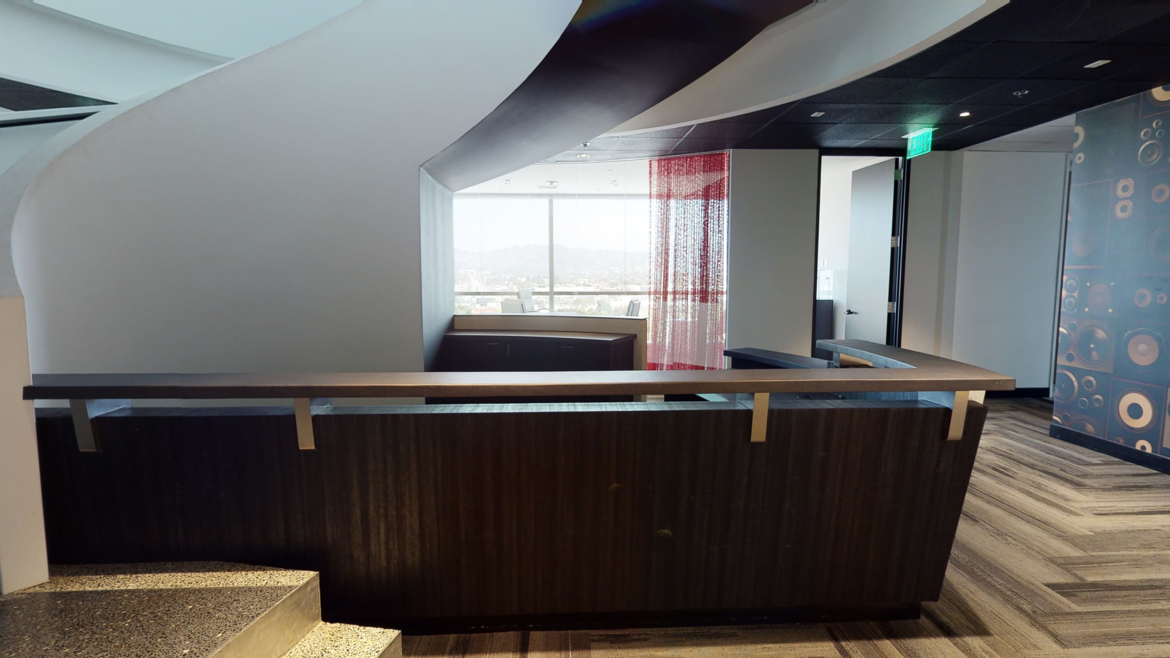 The image shows a modern office interior featuring a curved reception desk, large windows with a view, and a staircase leading to a different level.