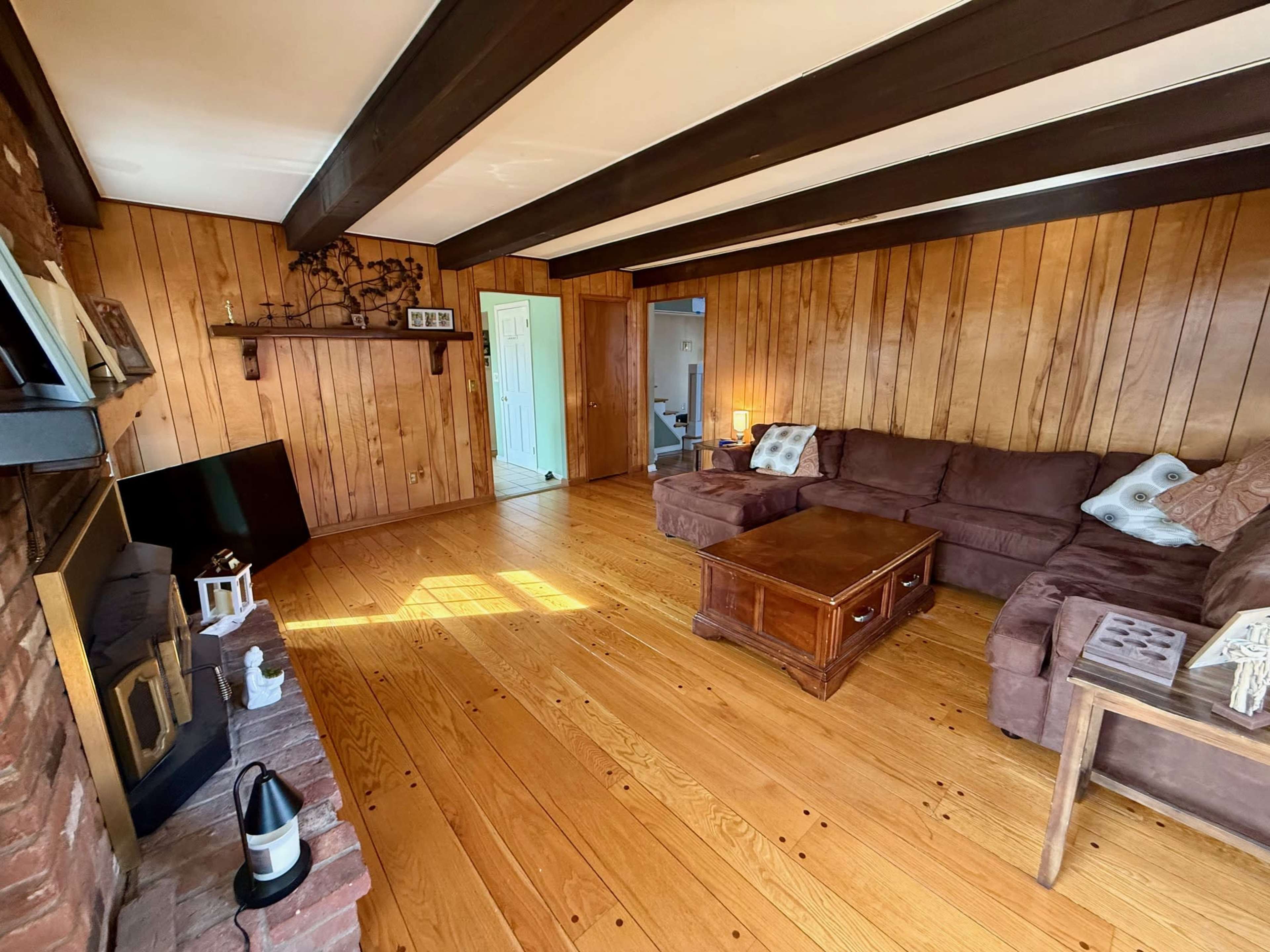 The image shows a spacious living room with wooden paneled walls, a brick fireplace, and a large sectional sofa arranged around a wooden coffee table.