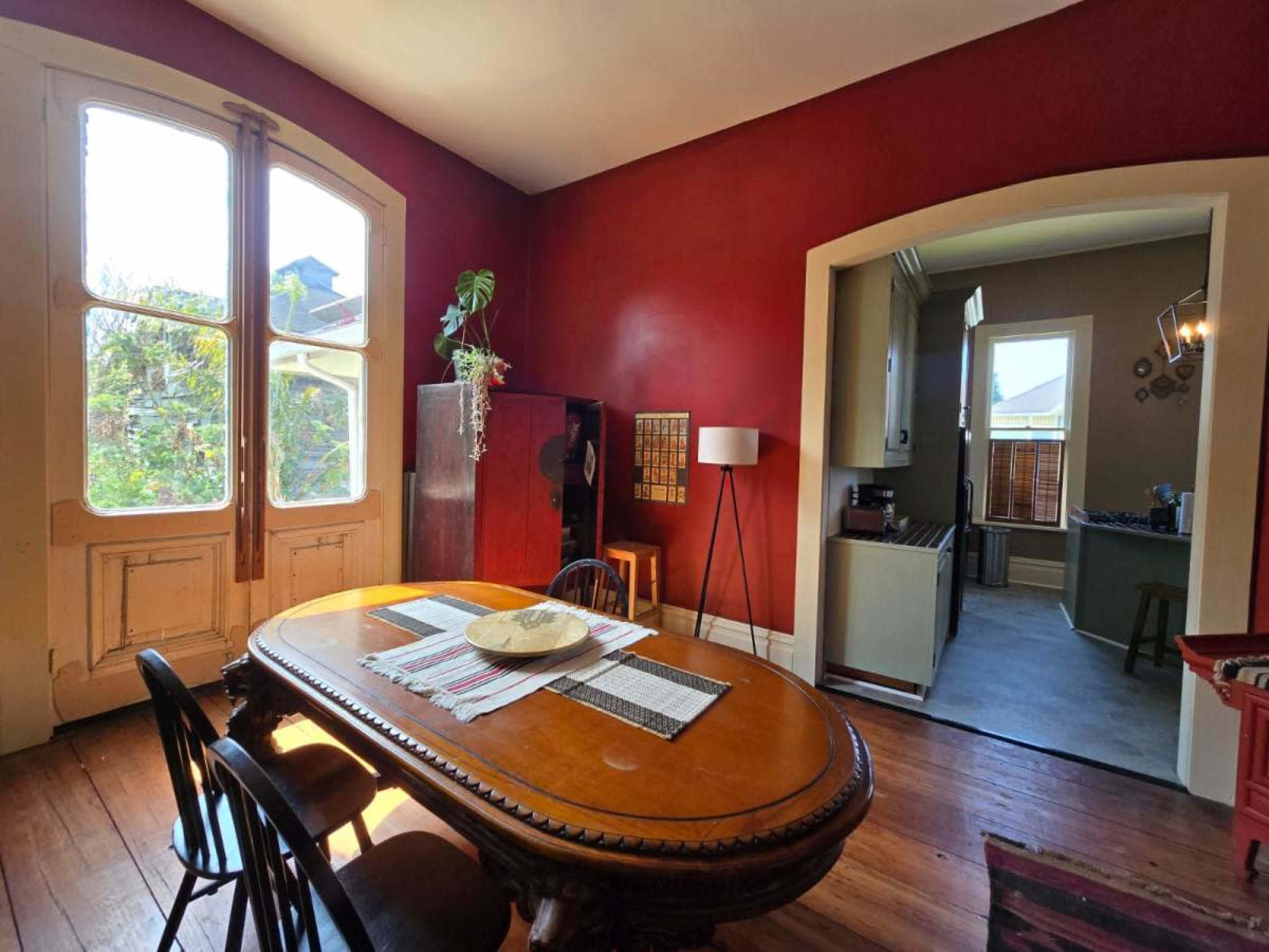 The image depicts a dining area with a wooden table and chairs, accompanied by large windows on one side and a doorway leading to another room on the opposite side, with red walls and wooden flooring.