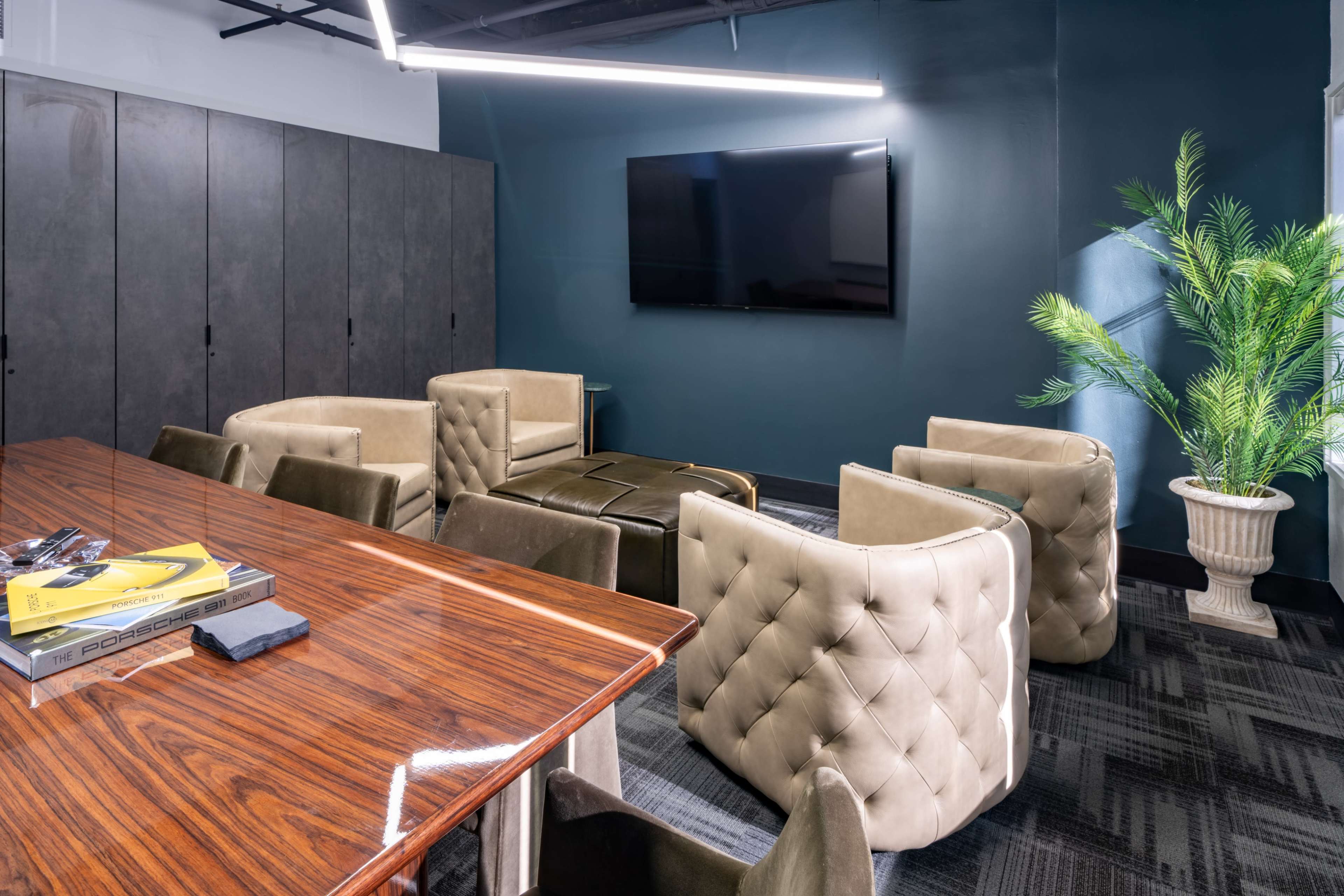 The image shows a modern meeting room with a polished wooden table, eight tufted chairs, a large television mounted on the wall, and a decorative plant in a corner.