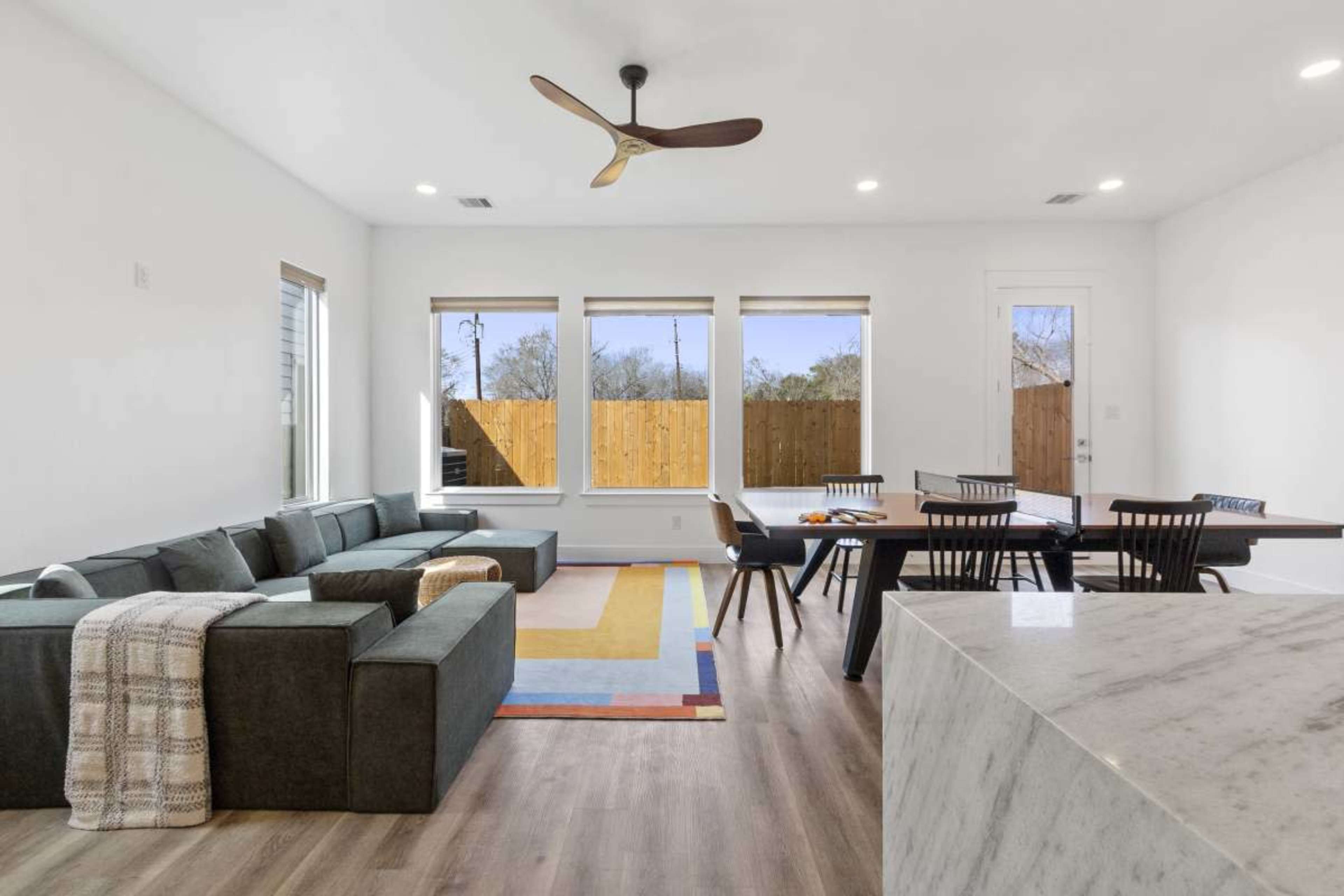 The image shows a modern living room with a sectional sofa, a dining area with a black table and chairs, large windows, and light-colored walls.