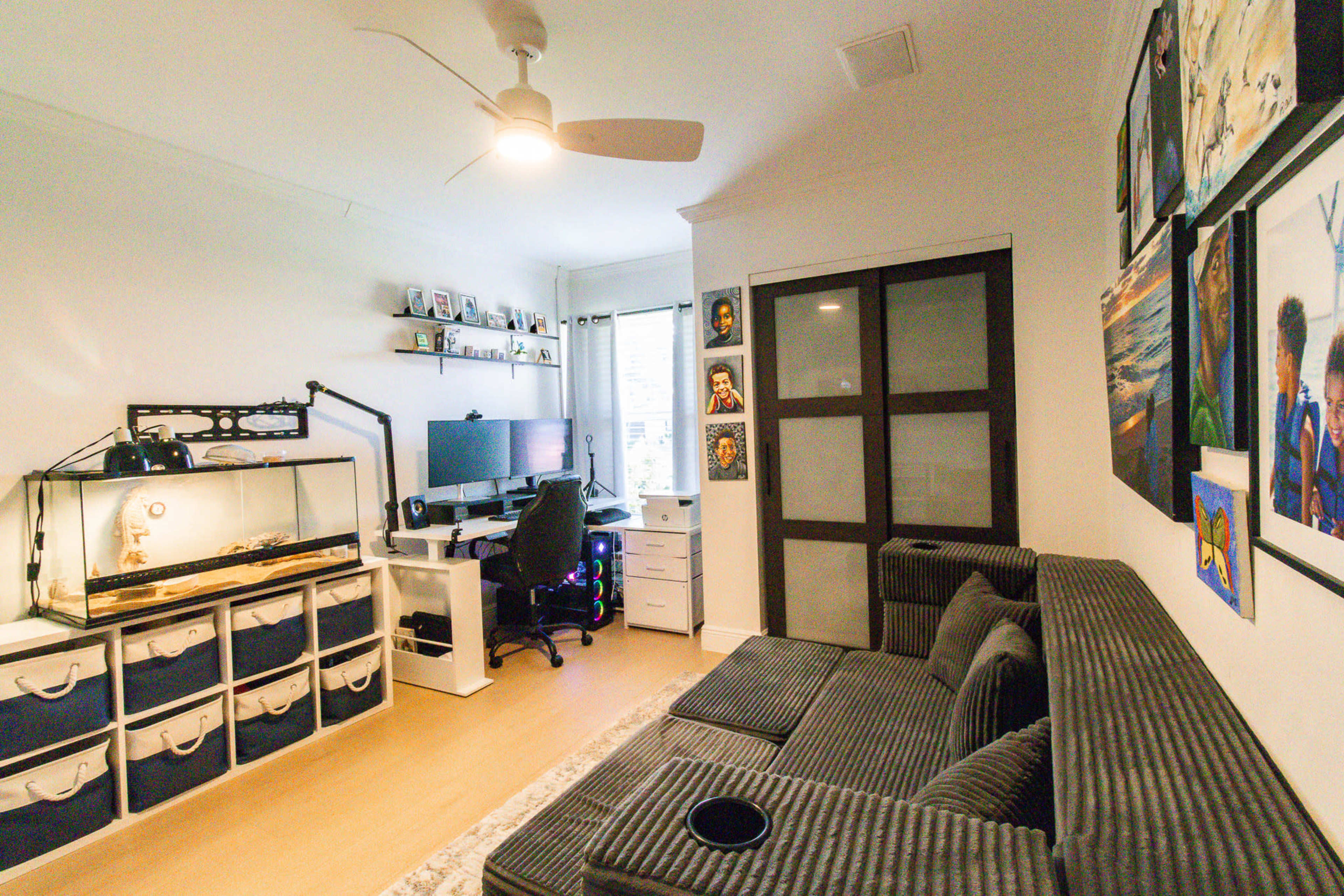 The image shows a modern home office setup with a computer desk, a comfortable gray sofa, a reptile tank, and artwork on the walls.