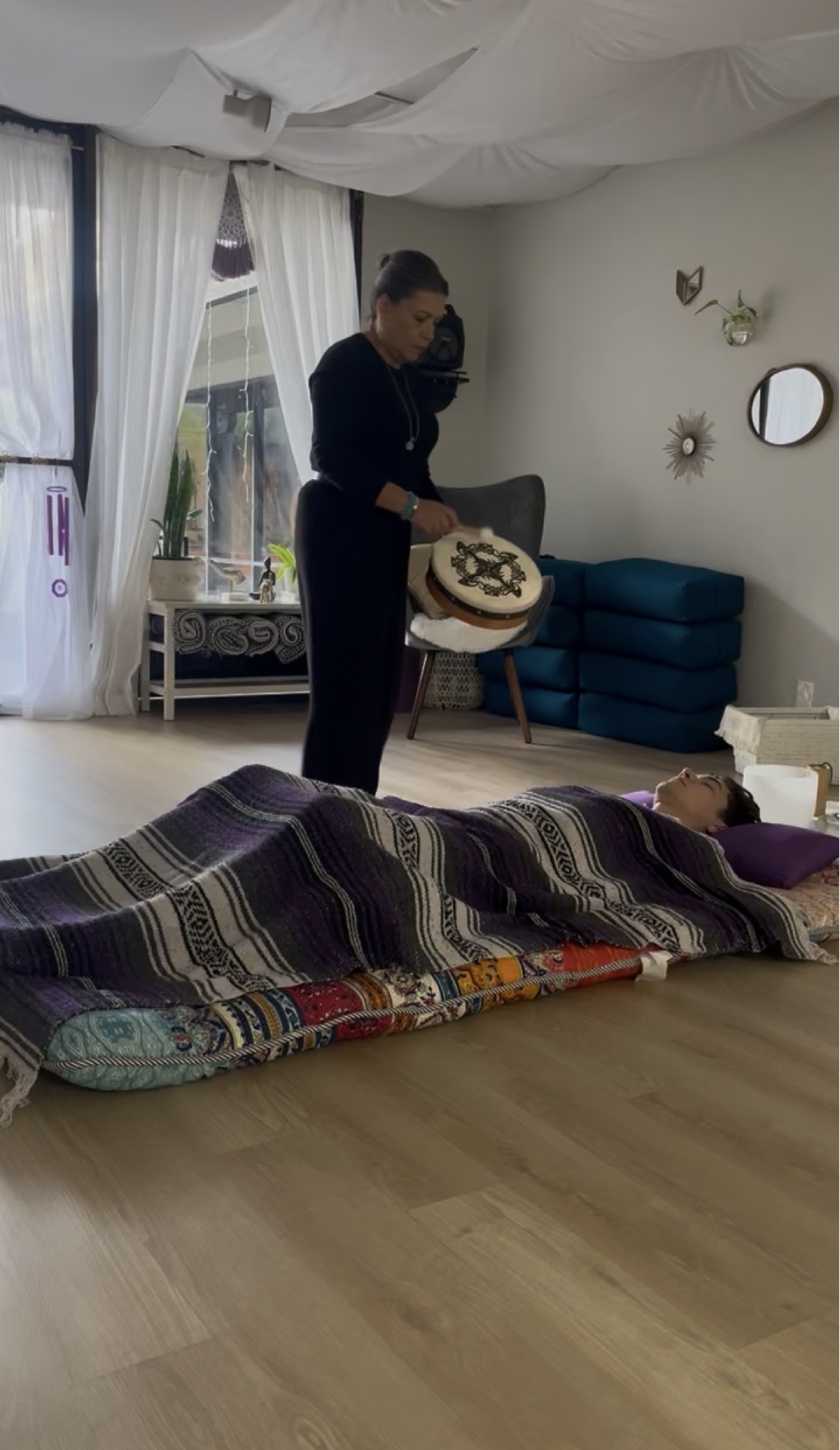 A woman stands beside a person lying under a blanket on the floor in a softly lit room, holding a drum.