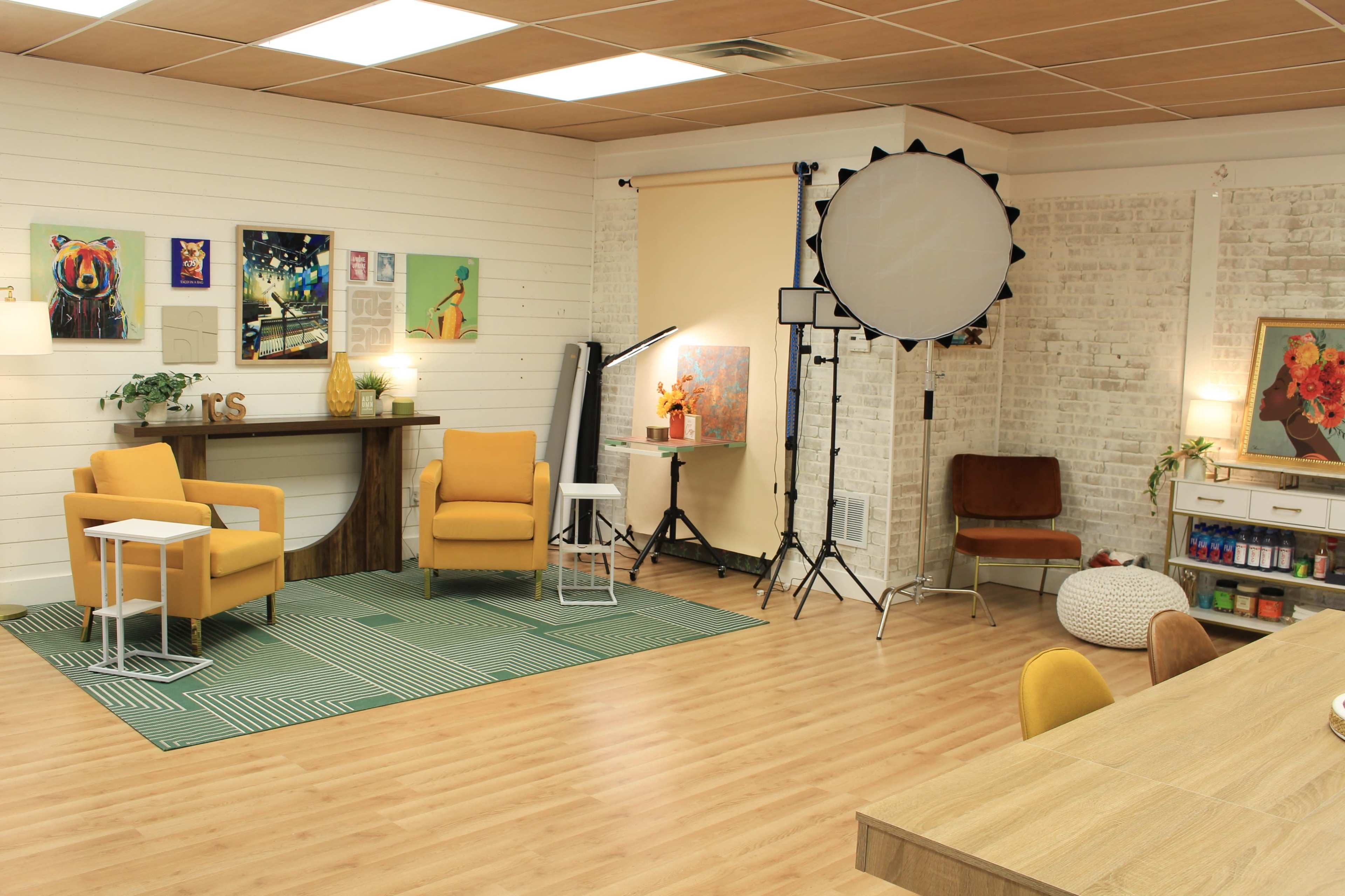 A bright studio space featuring two yellow armchairs, a green area rug, and various art pieces on the walls, along with photography equipment and a large round light umbrella.
