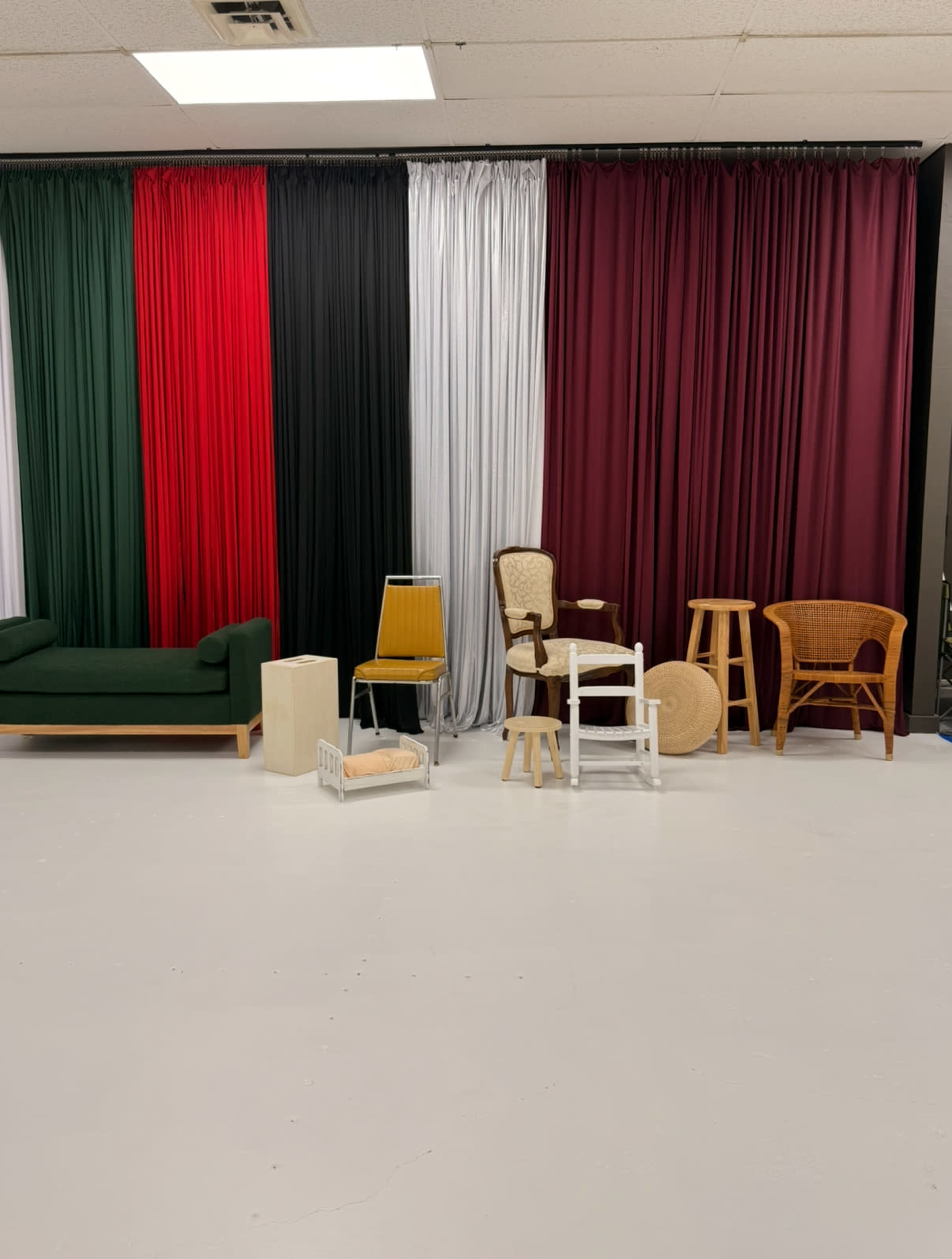 The image shows a studio space with a variety of furniture pieces in front of a backdrop of colorful fabric drapes in green, red, black, silver, and burgundy.