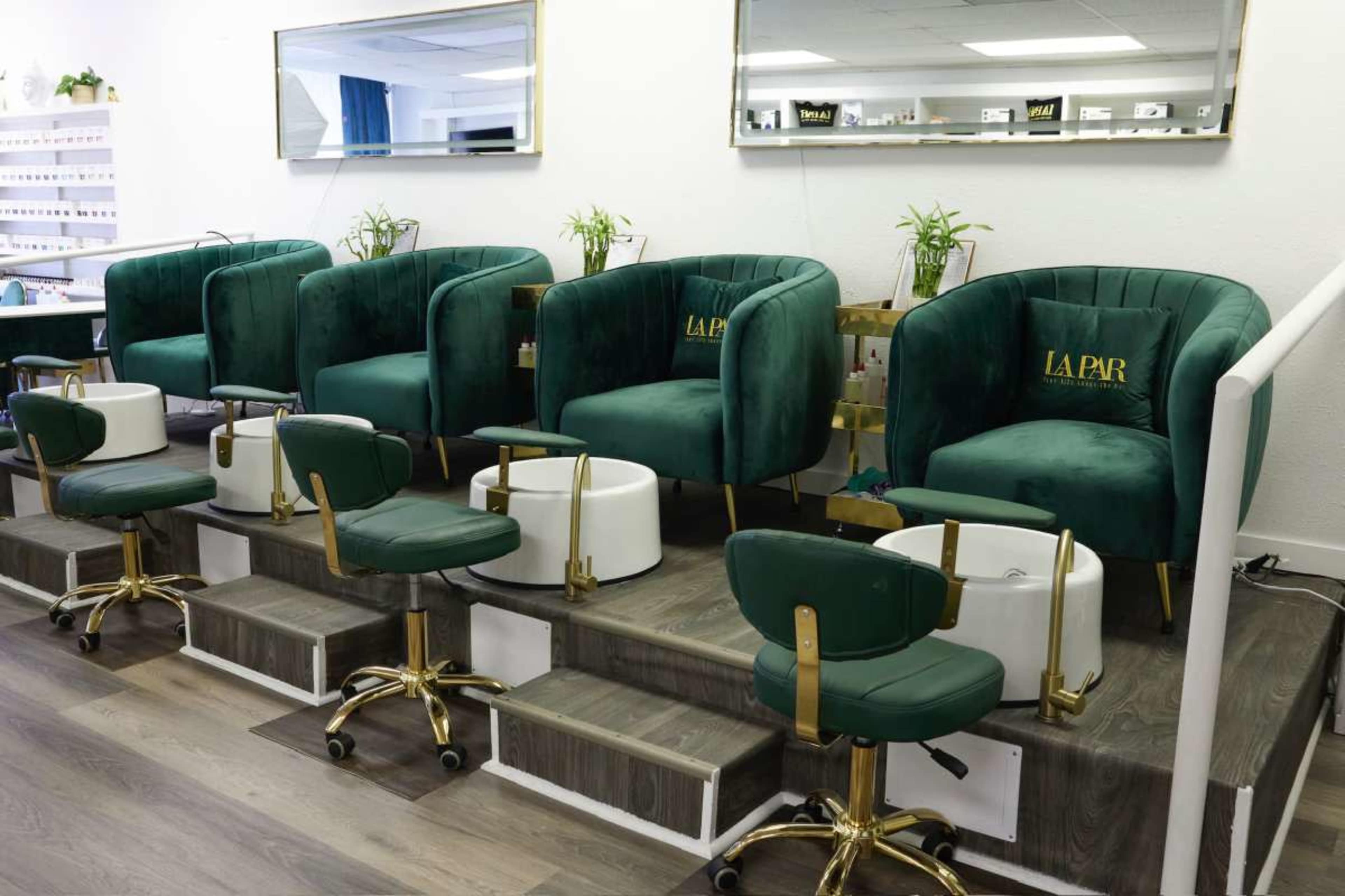 The image shows a nail salon interior featuring green velvet pedicure chairs with matching stools arranged in a row on a platform.