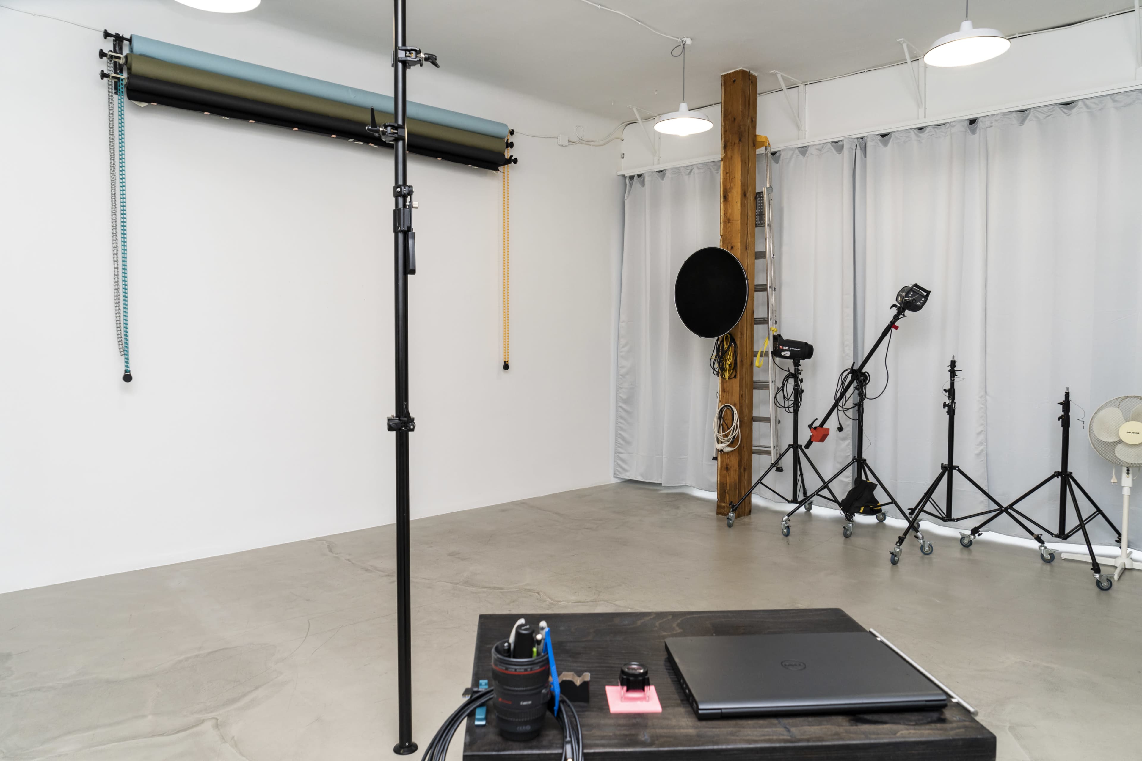 The image shows a spacious studio with a minimalist design, featuring a large empty wall, photography equipment, and a laptop on a table.