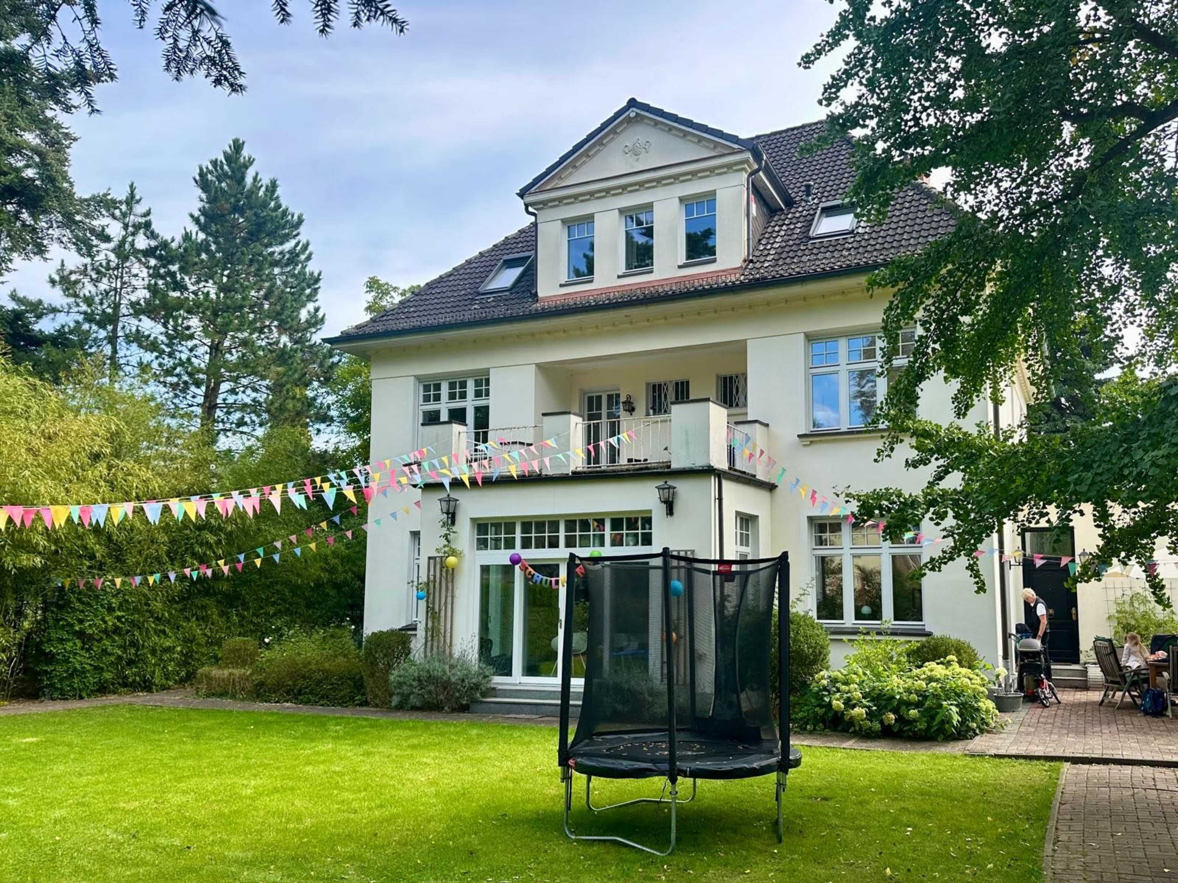 A large three-story house with a manicured lawn is decorated with colorful bunting and features a trampoline in the yard.