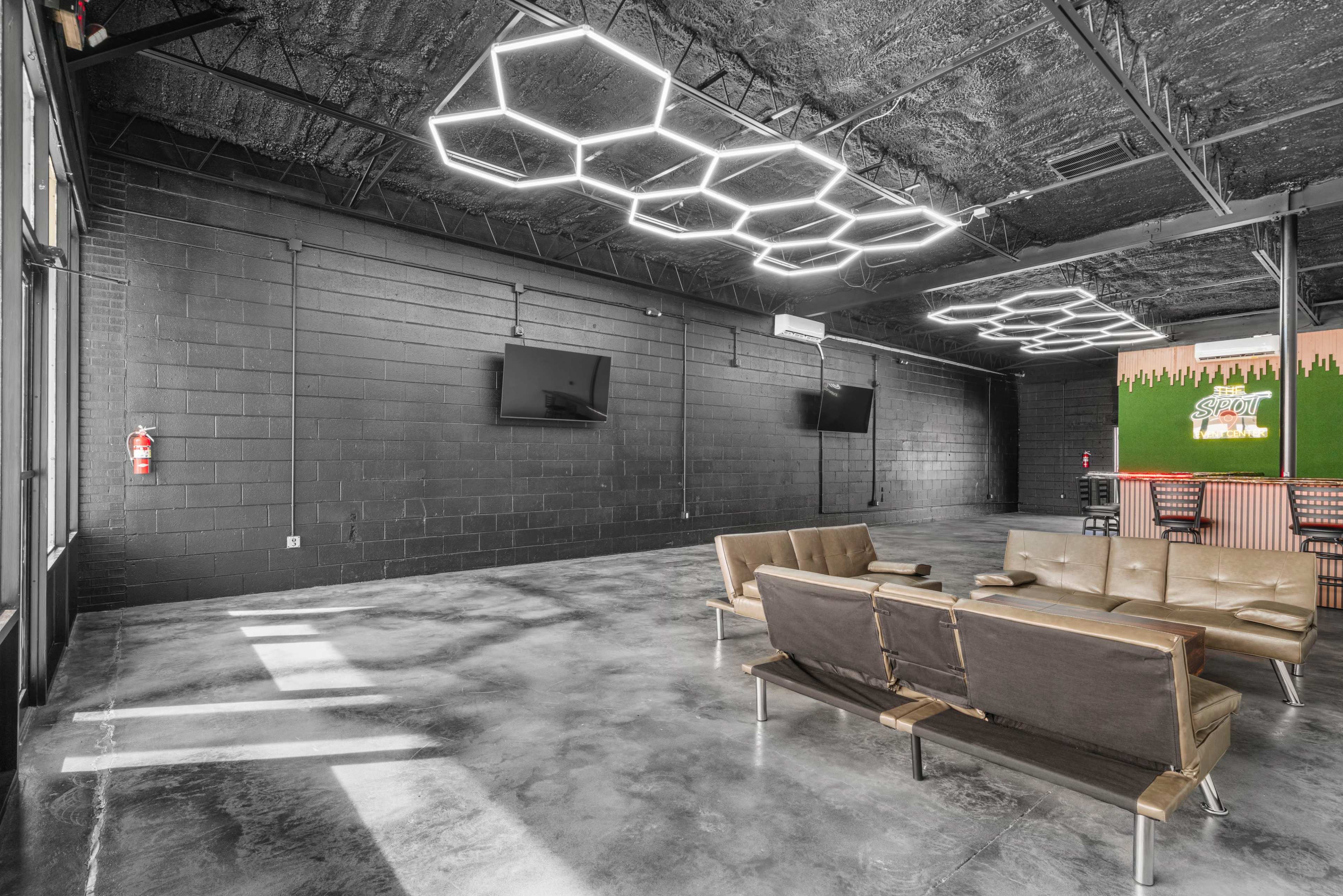 The image shows a large, open room with a polished concrete floor, black walls, and modern hexagon-shaped light fixtures on the ceiling, along with several flat-screen TVs mounted on the walls and a bar area in the background.