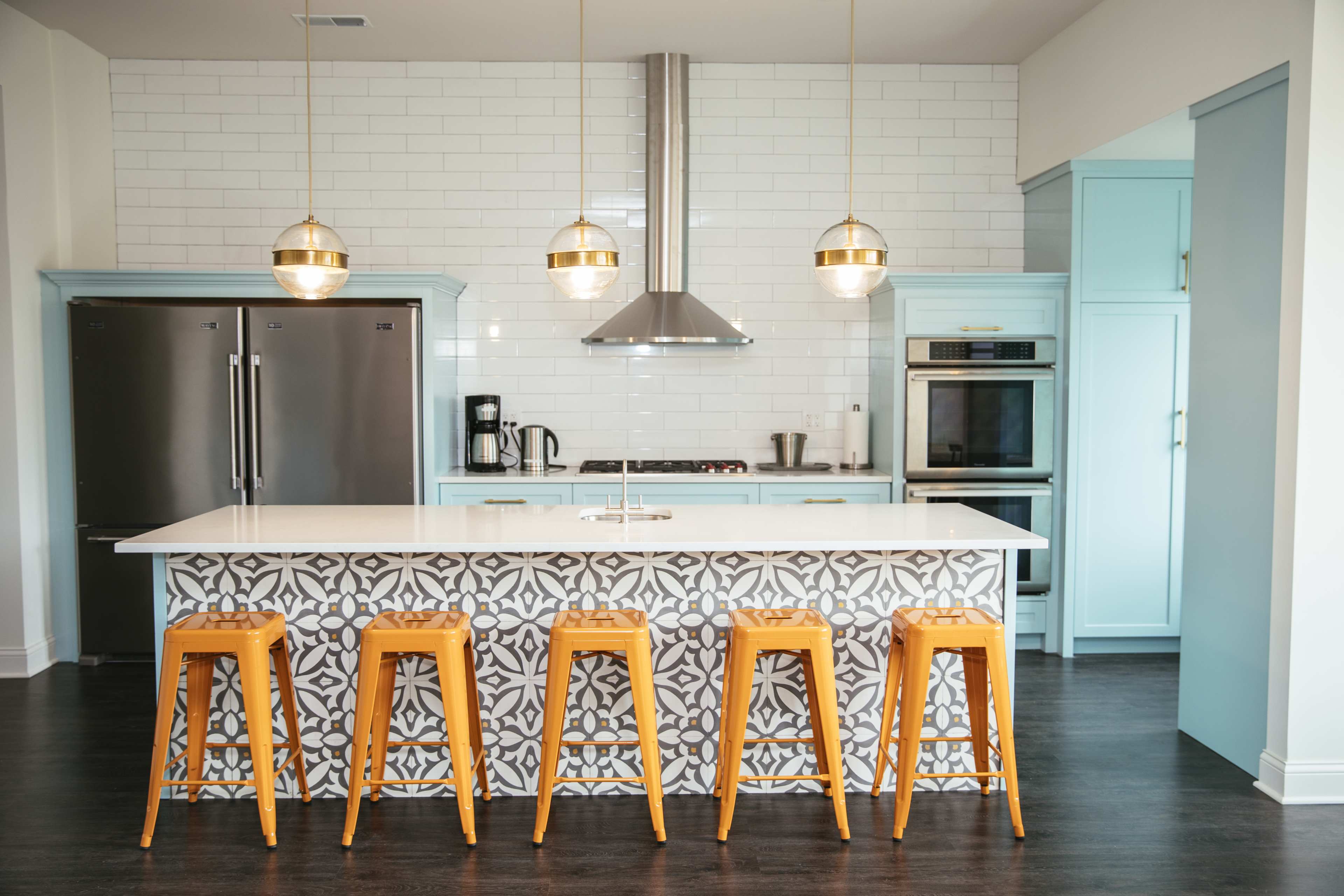 A modern kitchen features a large island with a patterned front, four yellow stools, stainless steel appliances, and pendant lights above.