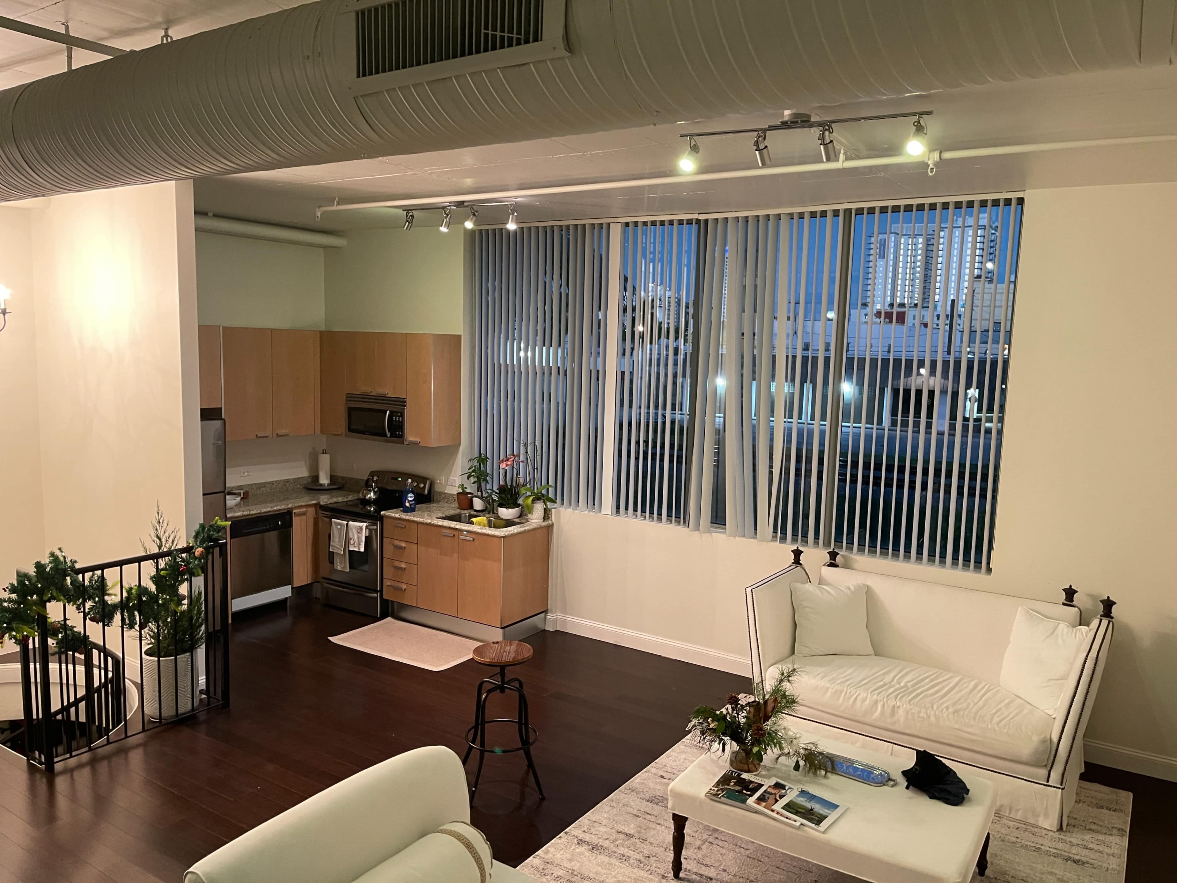 The image shows a modern loft-style living space with a kitchen, large windows, and a seating area featuring white couches and a coffee table.