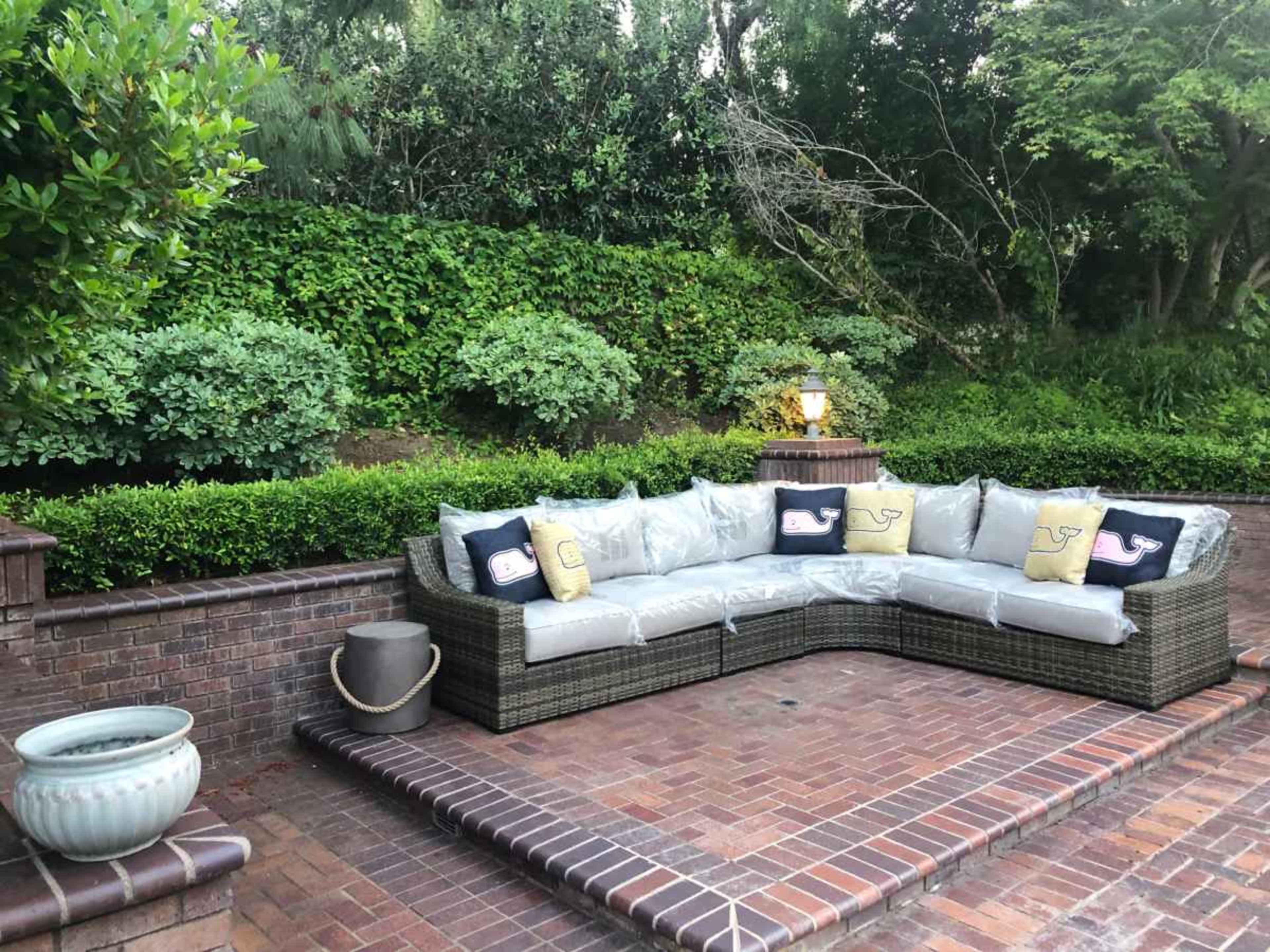 A sectional outdoor couch with cushions sits on a brick patio surrounded by green plants and shrubbery.