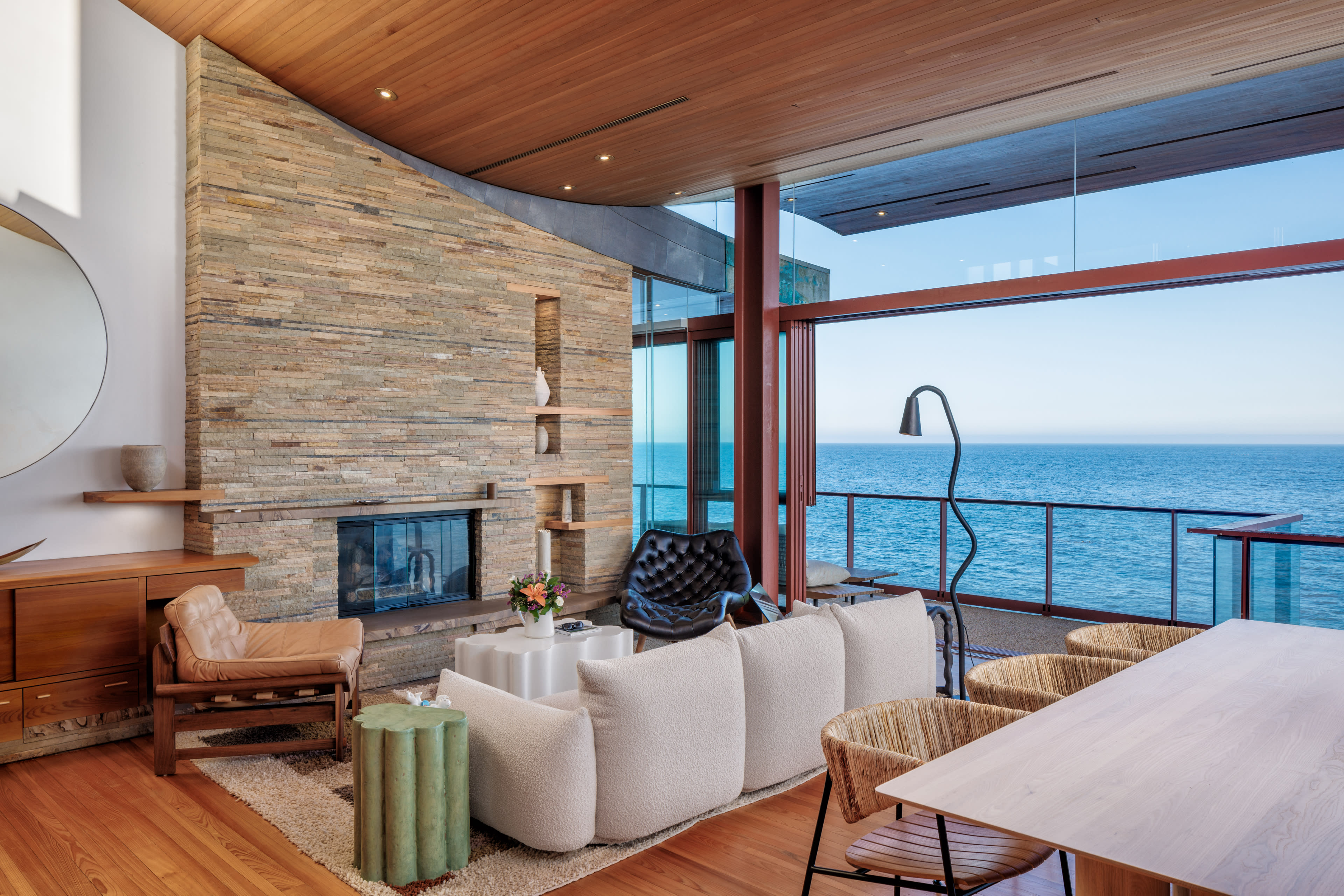 A spacious living room features a stone fireplace, large windows overlooking the ocean, and modern furniture arranged for relaxation.