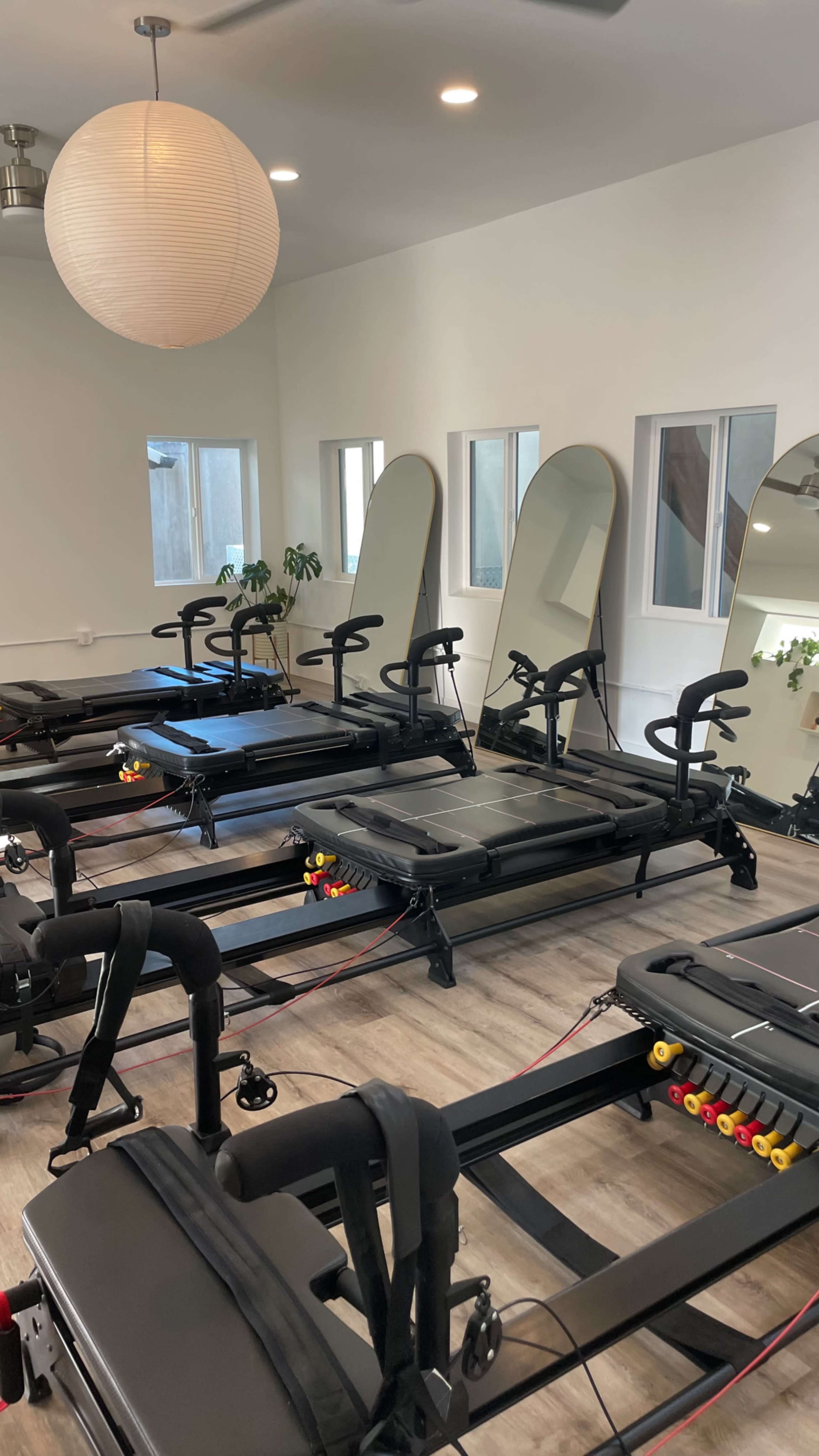 The image shows a fitness studio equipped with several reformer Pilates machines, mirrors, and large windows letting in natural light.