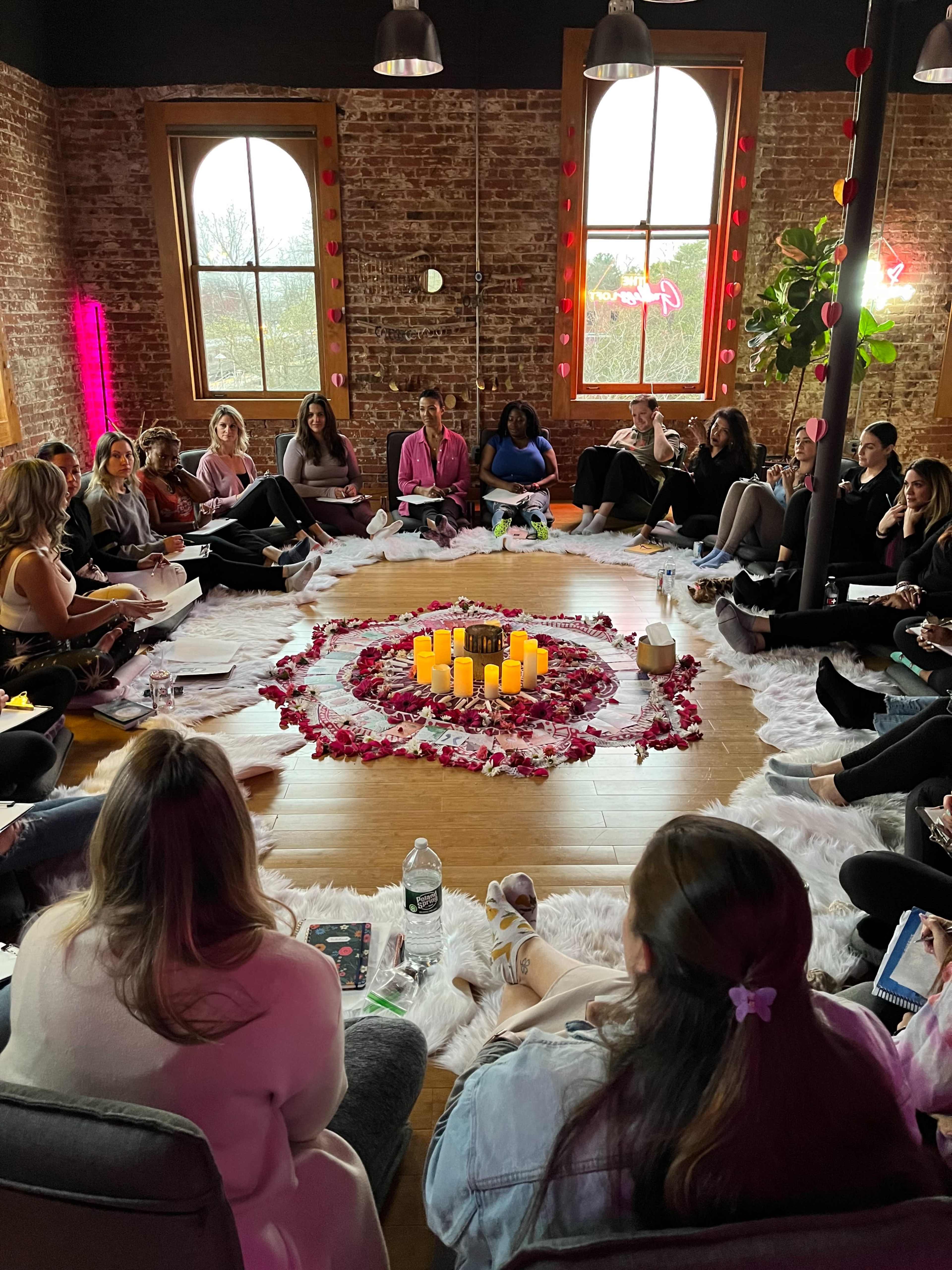 A group of women sits in a circular arrangement on the floor around a centerpiece of candles and flower petals in a warmly lit room with brick walls.