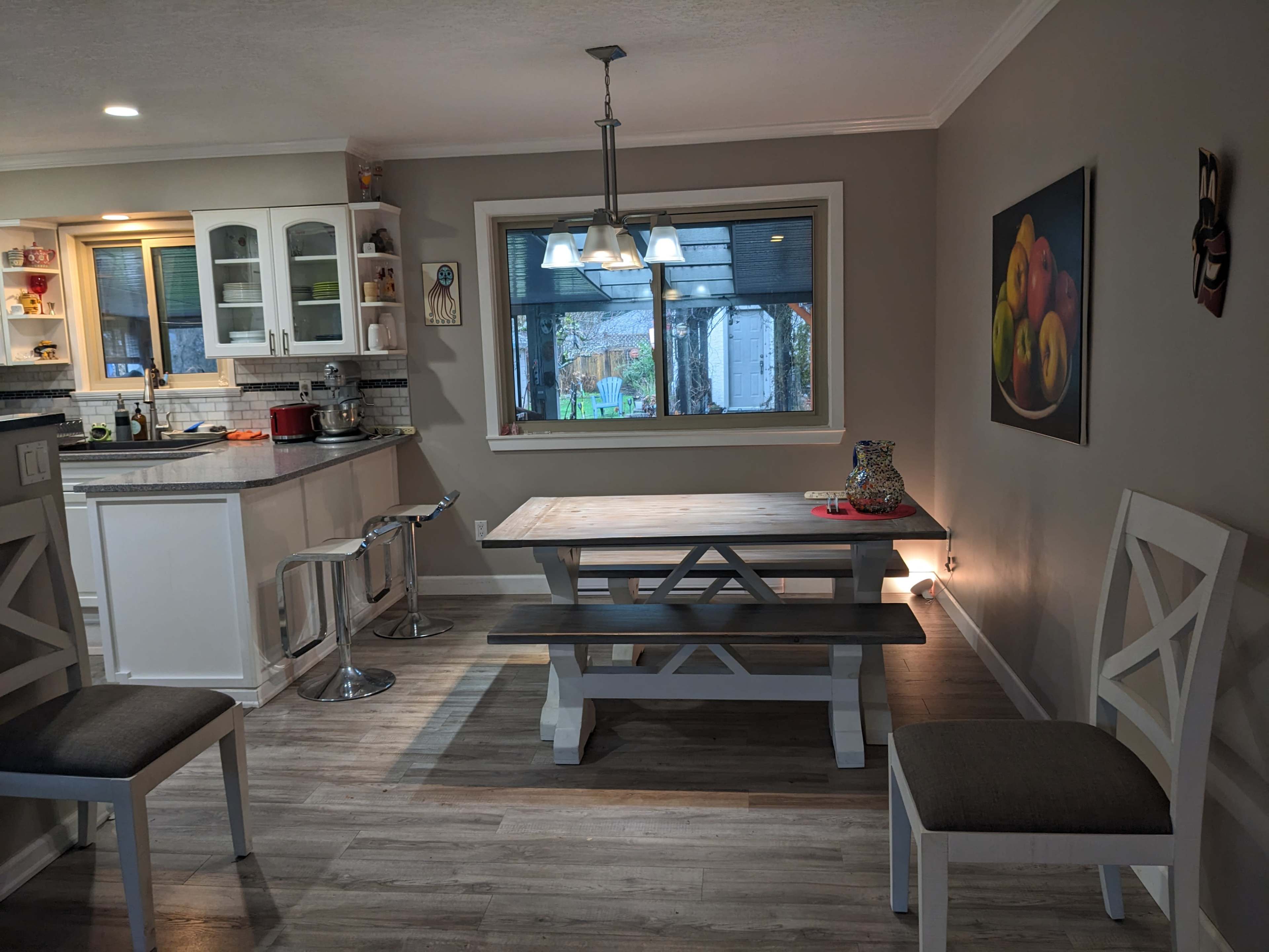 The image shows a modern kitchen-dining area with a wooden table, bench seating, and a window overlooking a backyard, illuminated by overhead lights.