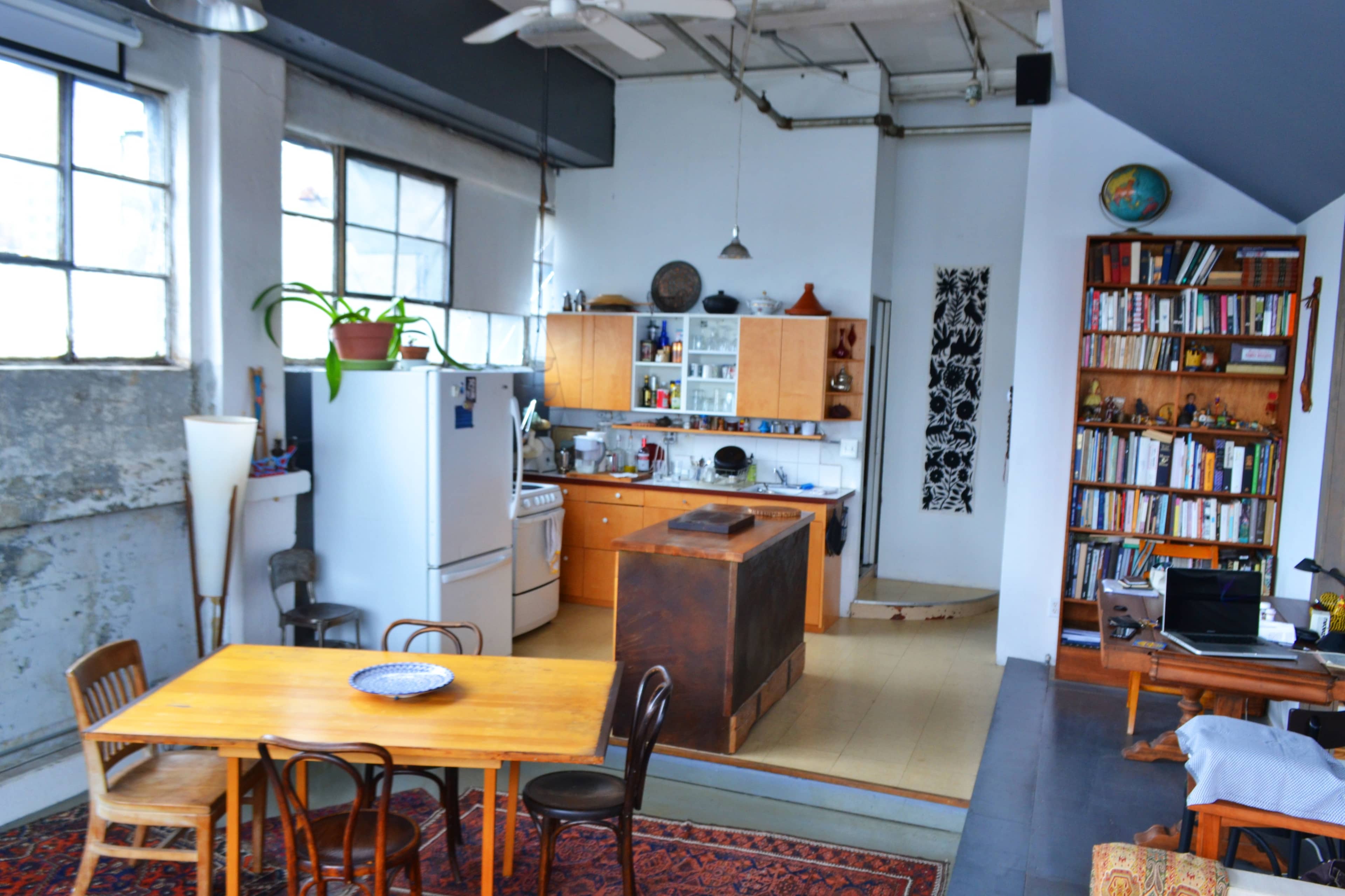 A spacious kitchen and dining area featuring a wooden table, chairs, and a refrigerator, with shelves and cabinets filled with dishes and books.