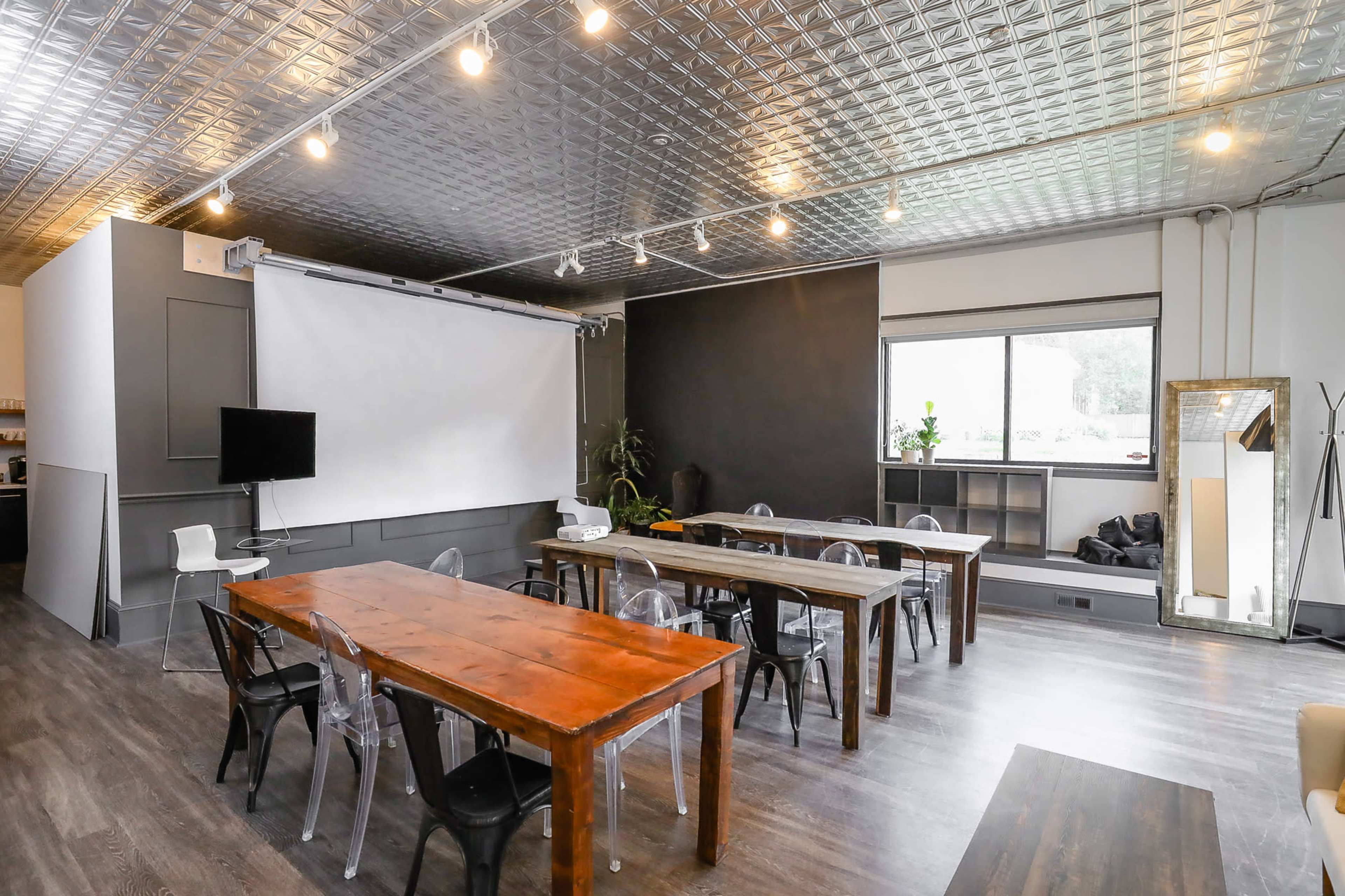 The image shows a modern meeting room with two long wooden tables, transparent chairs, a projector screen, and a TV on the wall.