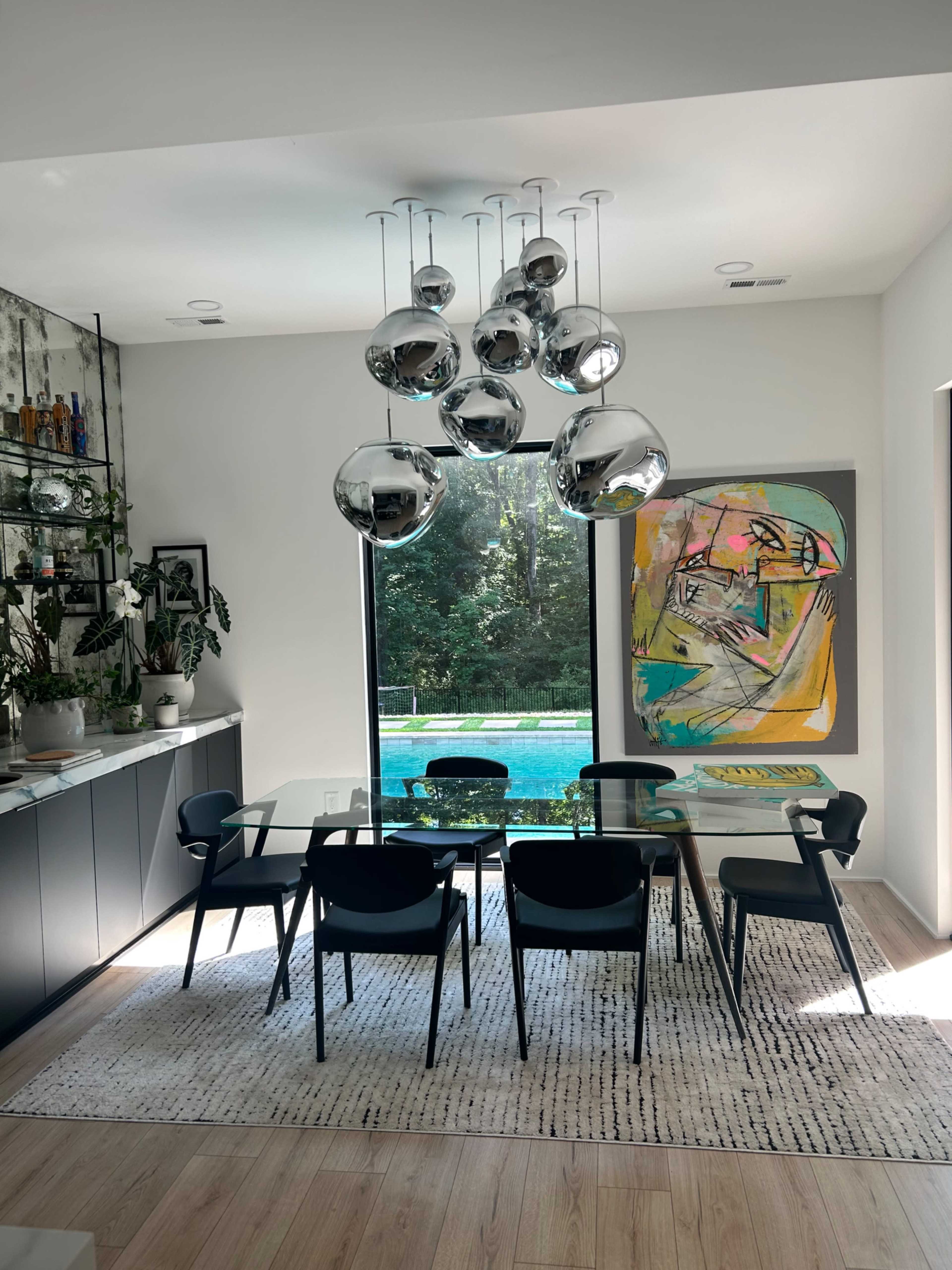 A modern dining area features a glass table surrounded by black chairs, a colorful abstract painting on the wall, and a view of a pool through large windows.