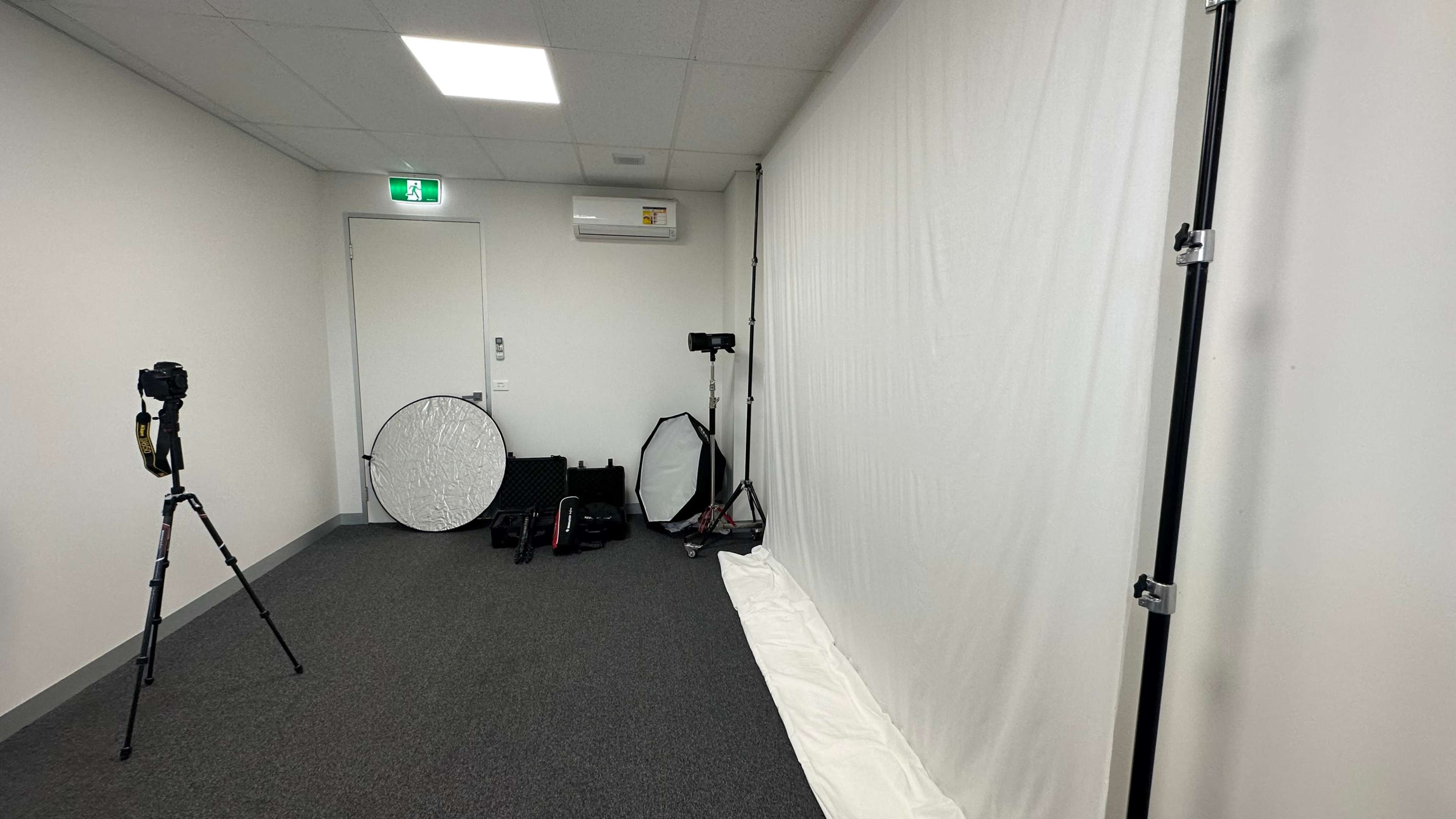 A photography studio with a white backdrop, a tripod-mounted camera, lighting equipment, and various photography accessories arranged along the wall.