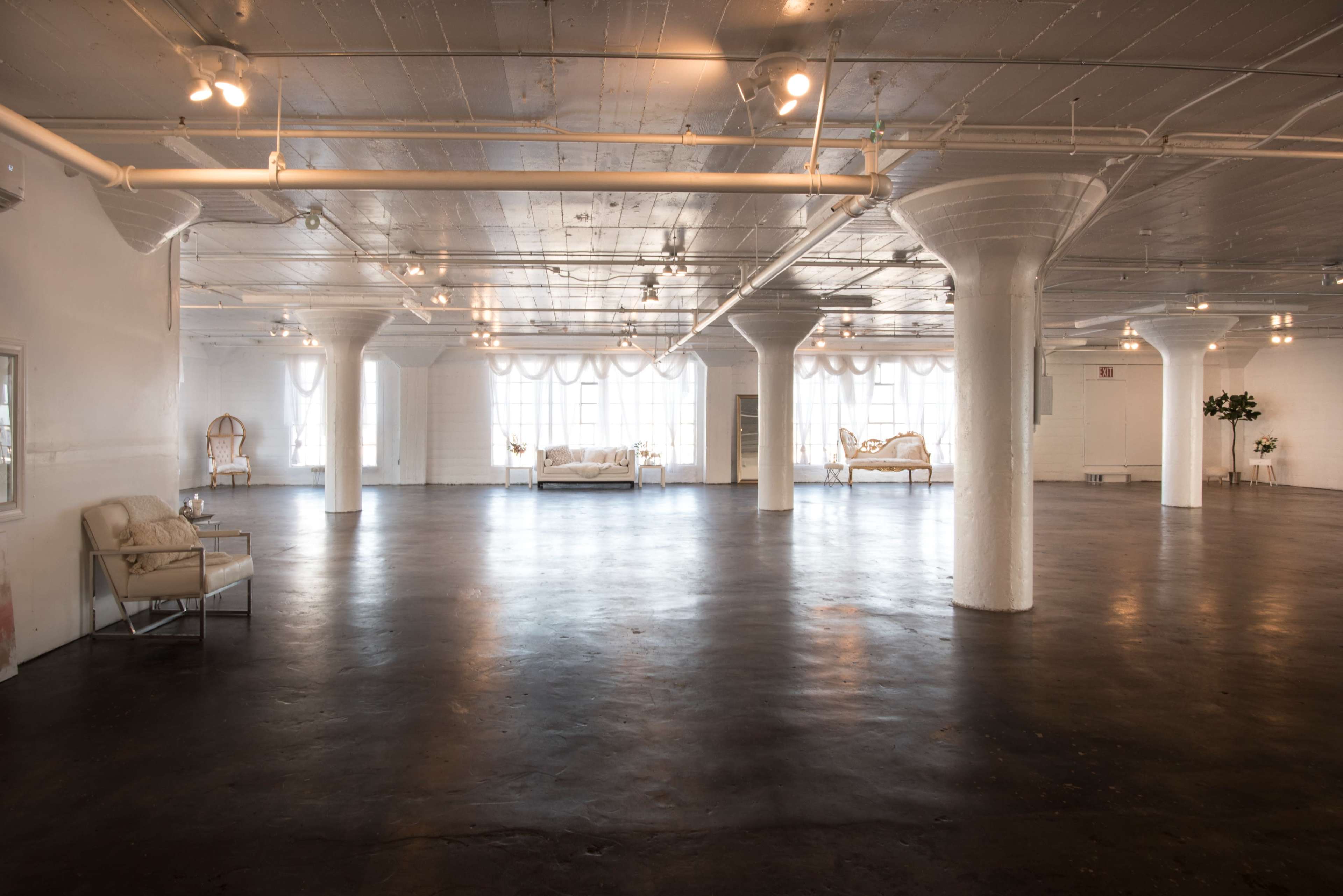 A spacious, well-lit interior with white walls and large windows, featuring minimalist furnishings and polished concrete flooring.